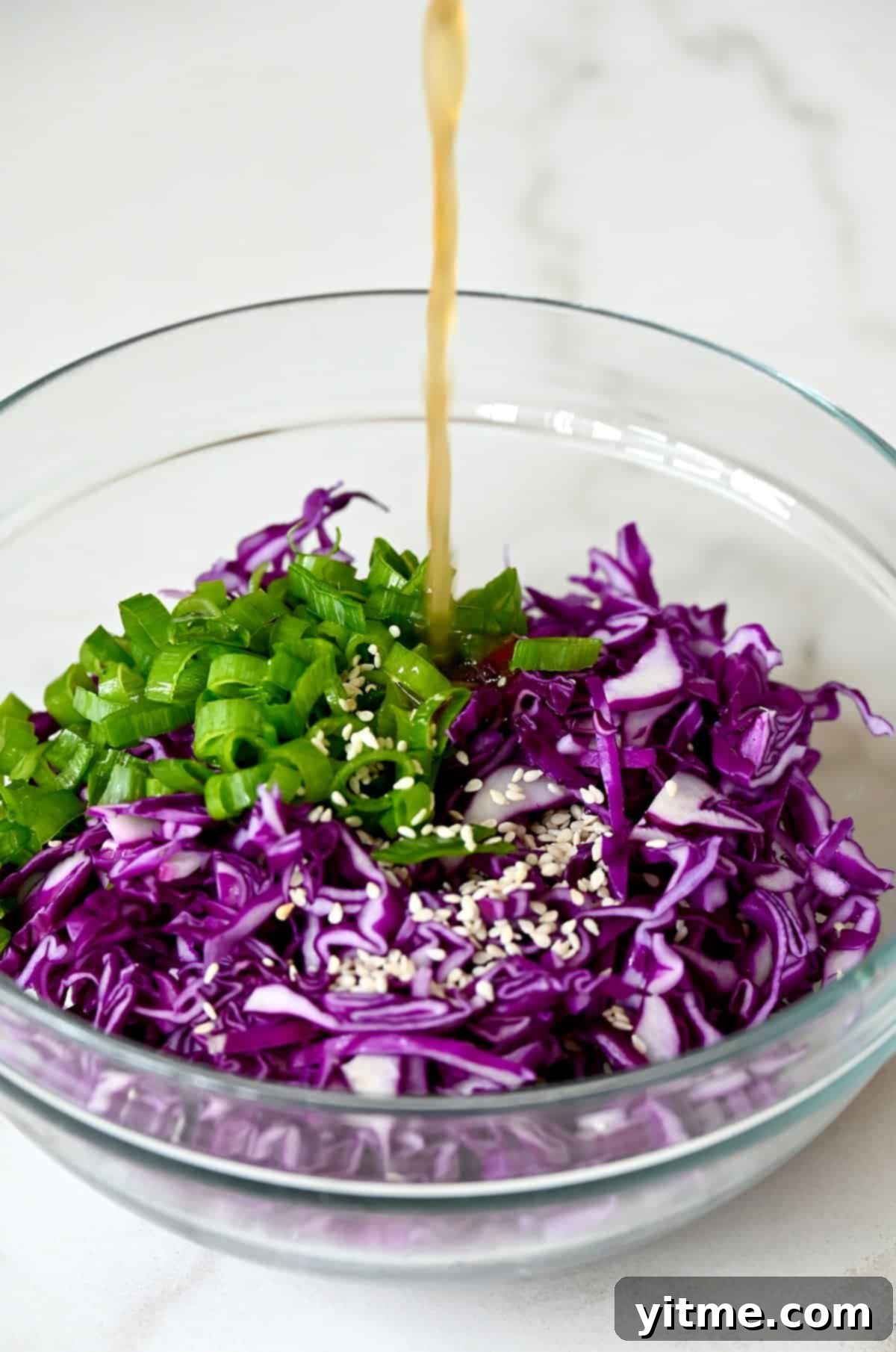Close-up of Asian dressing being generously poured over a colorful mix of shredded purple cabbage, green scallions, and white sesame seeds in a large mixing bowl.