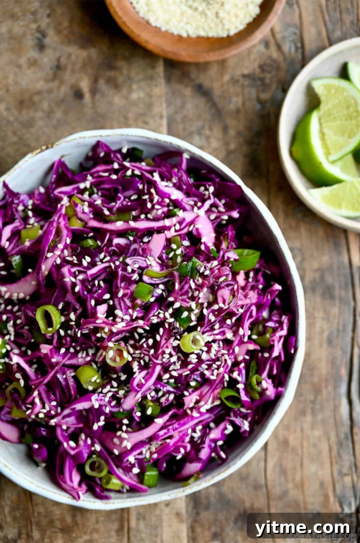 Large white salad bowl brimming with colorful Asian slaw, garnished with extra sesame seeds, accompanied by a small bowl of fresh lime wedges, ready for serving.