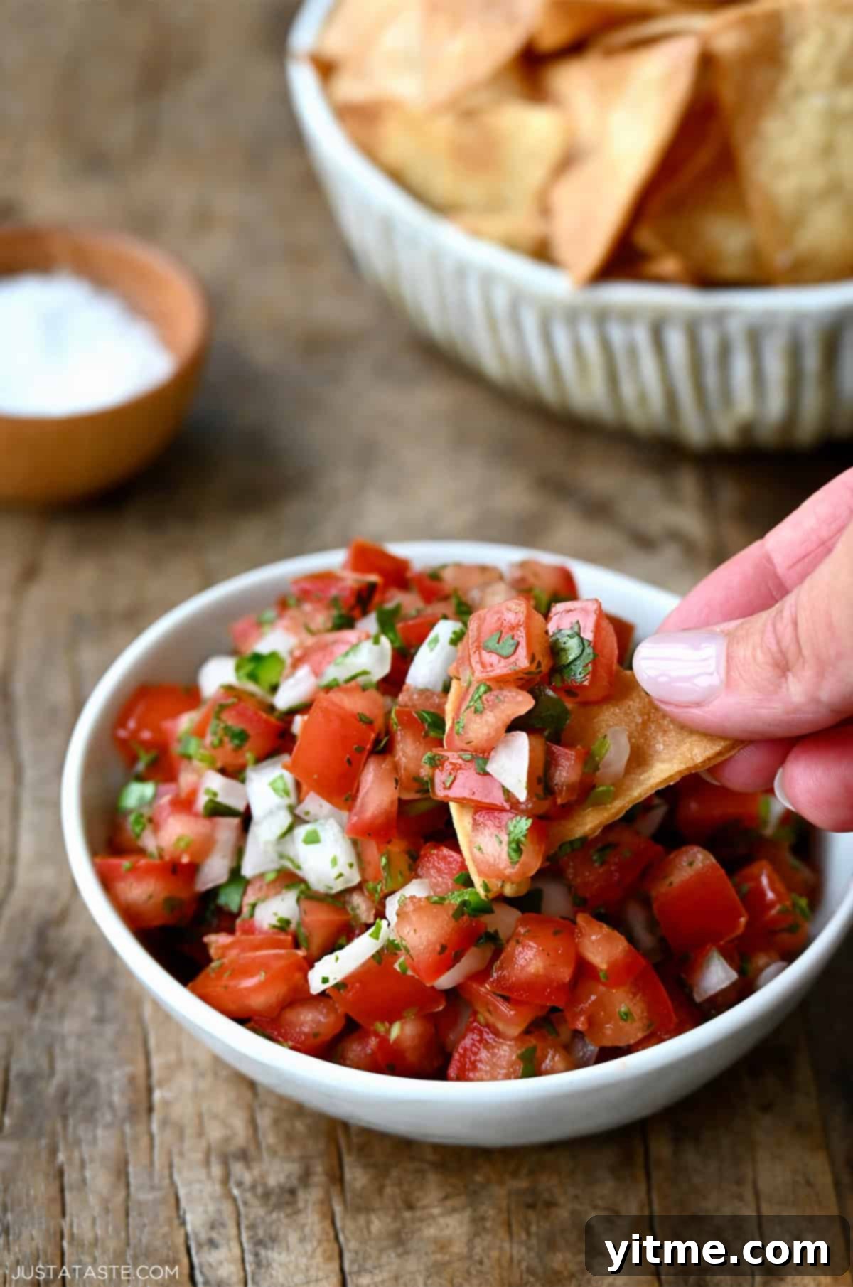A hand holding a tortilla chip scoops up homemade pico de gallo from a small white bowl.