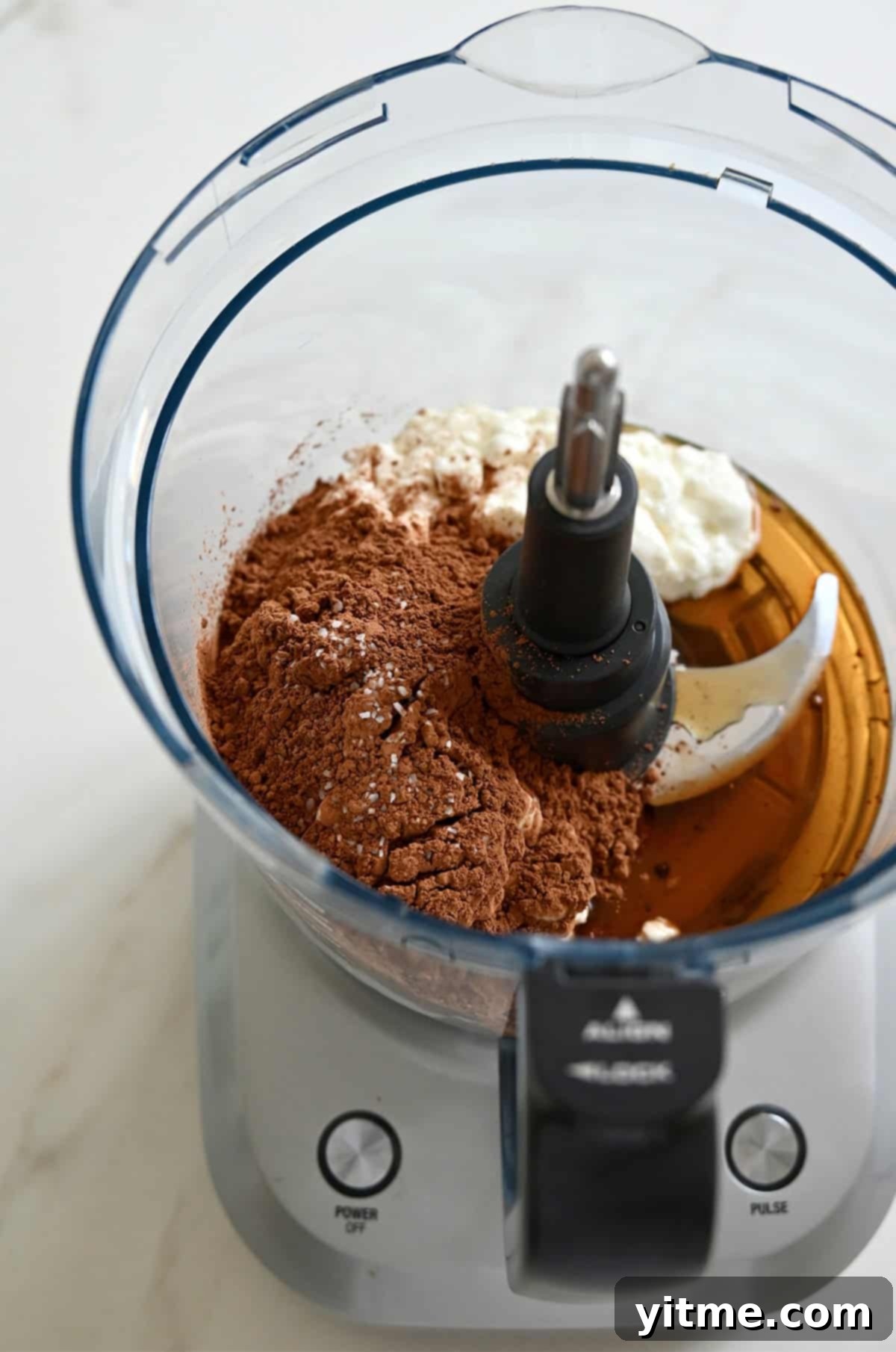 A food processor containing cottage cheese, cocoa powder, maple syrup and vanilla extract.