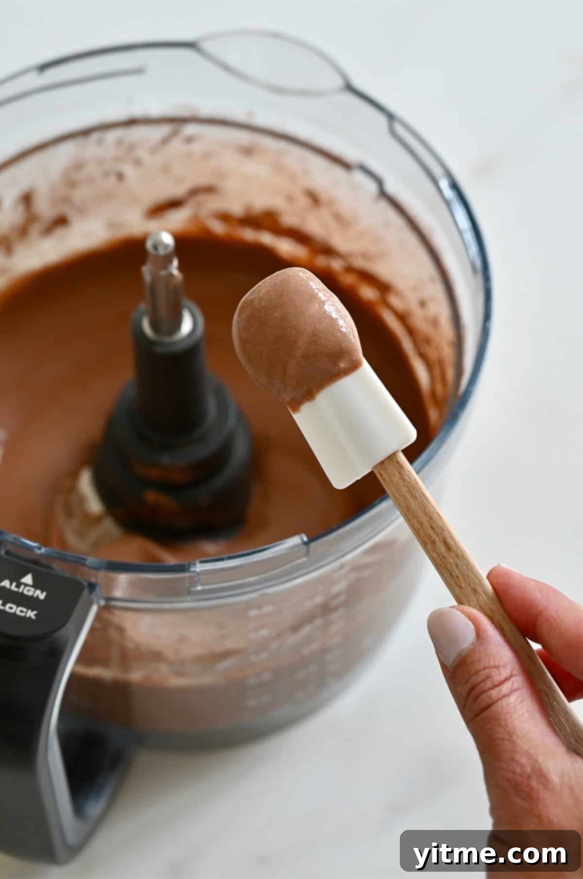 A white spatula with mousse on it with a food processor bowl in the background.
