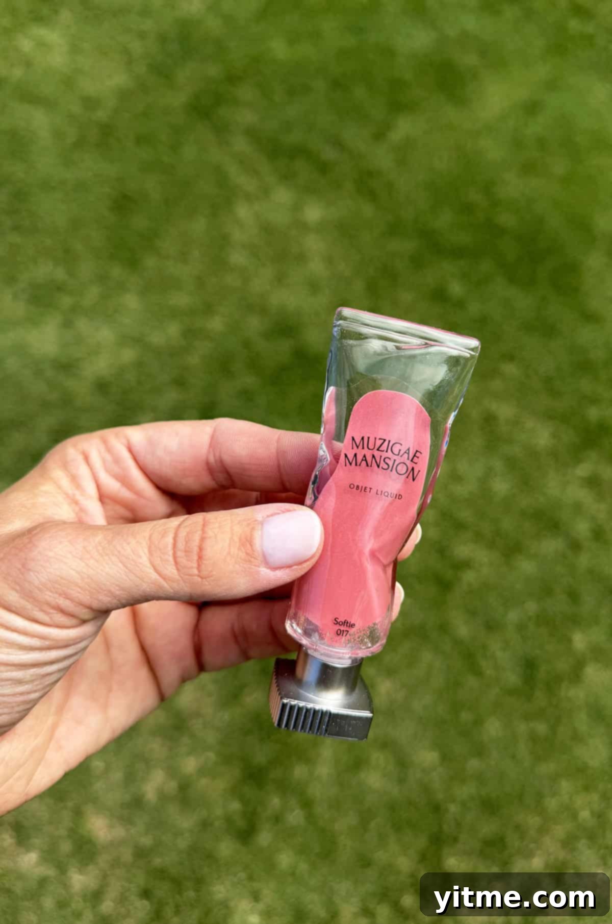 Kelly's Monthly Edit: July 2025 6 A delicate hand holding a chic pink tube of Muzigae Mansion lipstick against a soft green, out-of-focus grassy background, highlighting the product's elegant design.