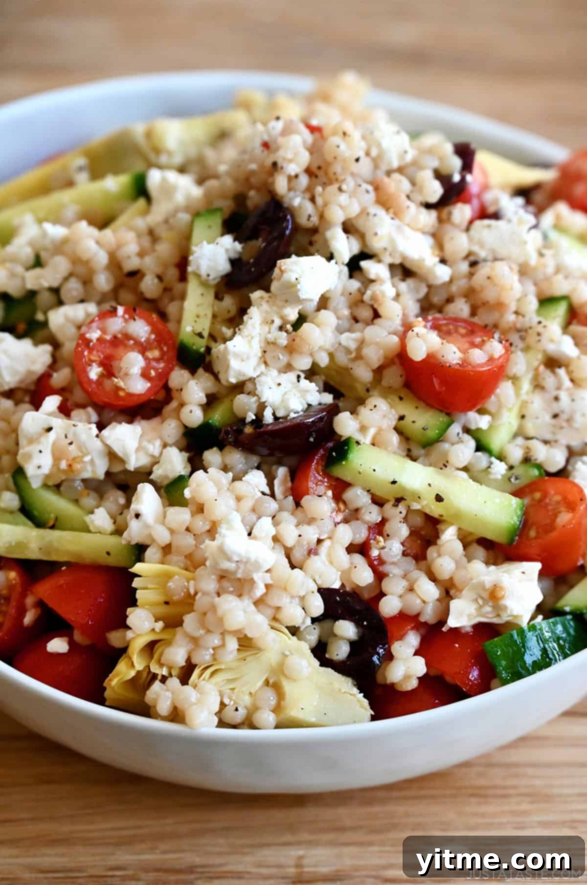 A beautifully presented Mediterranean couscous salad with a zesty red wine vinaigrette, fresh cucumbers, sweet tomatoes, briny Kalamata olives, and creamy crumbled feta cheese, elegantly arranged in a large white serving bowl.