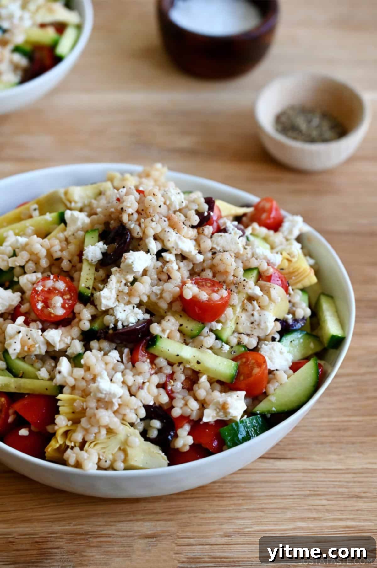 A large serving bowl showcasing a vibrant Mediterranean couscous salad, featuring pearl couscous, sliced cucumbers, cherry tomatoes, Kalamata olives, artichoke hearts, and crumbled feta cheese, all glistening with a red wine vinegar and lemon juice vinaigrette.