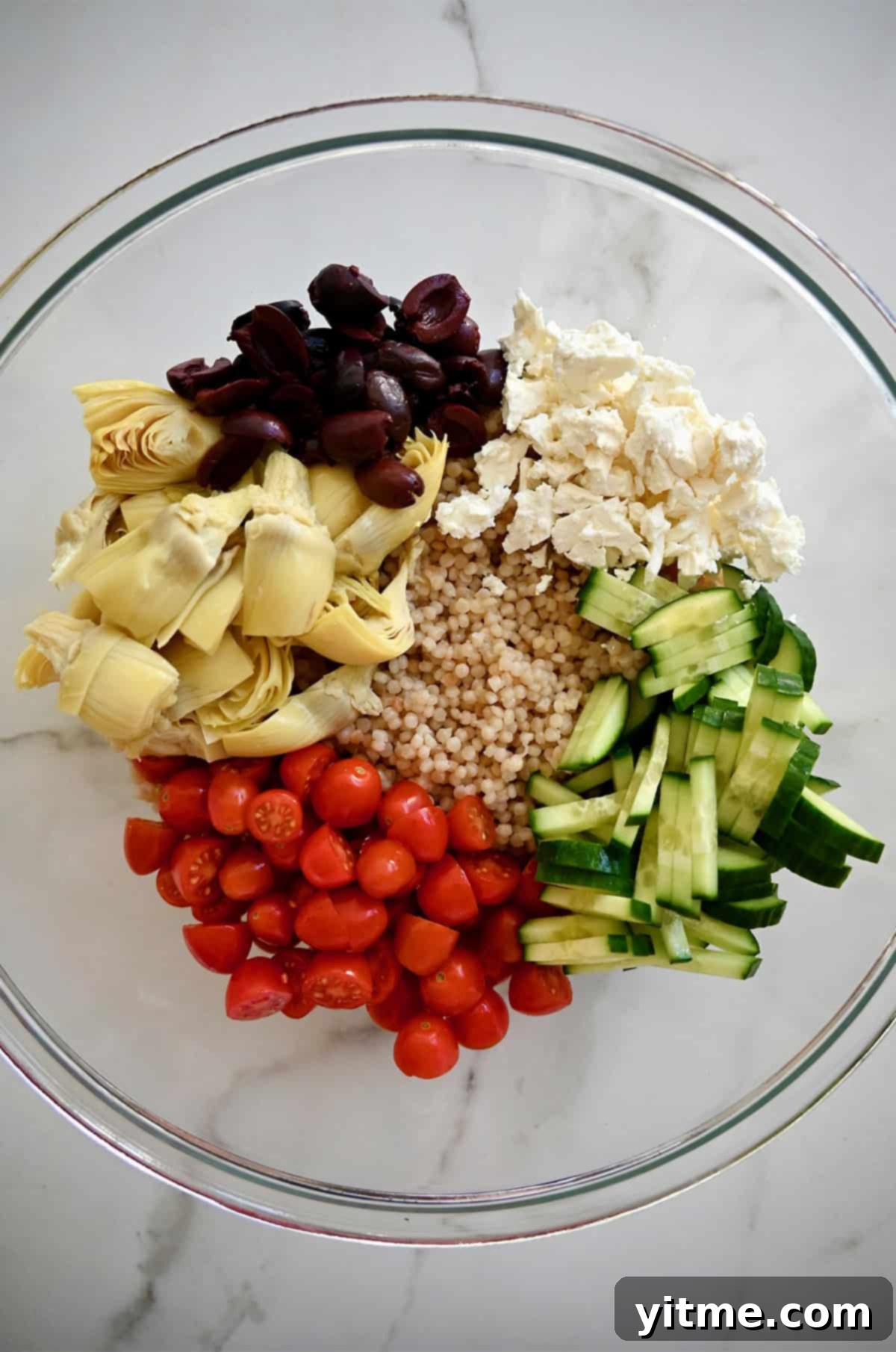 A large white bowl showcasing a vibrant Mediterranean couscous salad, featuring cooked pearl couscous mixed with halved cherry tomatoes, artichoke hearts, sliced Kalamata olives, crumbled feta cheese, and crisp sliced cucumbers, ready for serving.