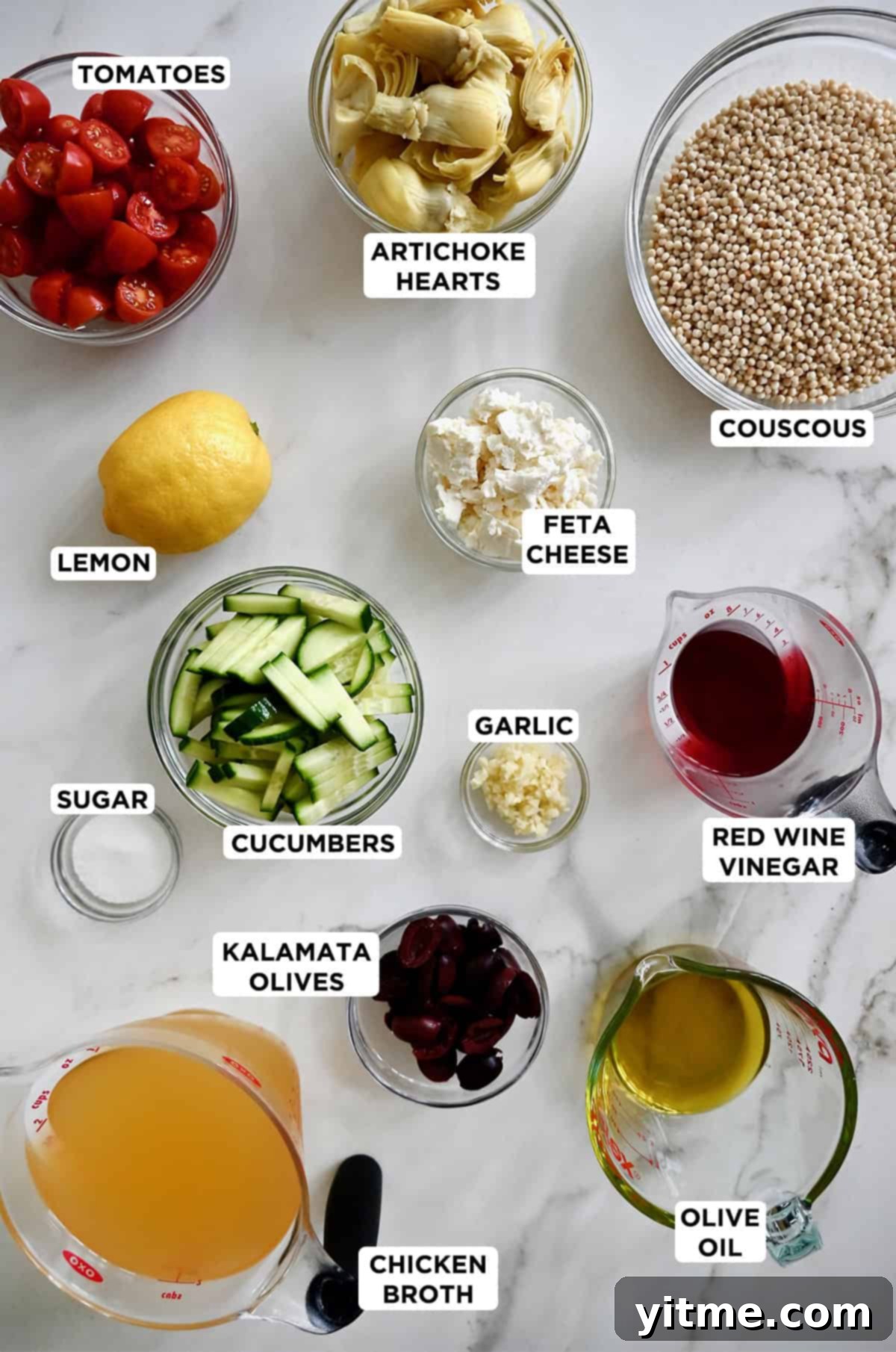 A collection of fresh ingredients laid out, including pearl couscous, cherry tomatoes, cucumbers, Kalamata olives, a can of artichoke hearts, a block of feta cheese, and bottles of olive oil and red wine vinegar, ready to make a Mediterranean couscous salad with a tangy red wine vinaigrette.