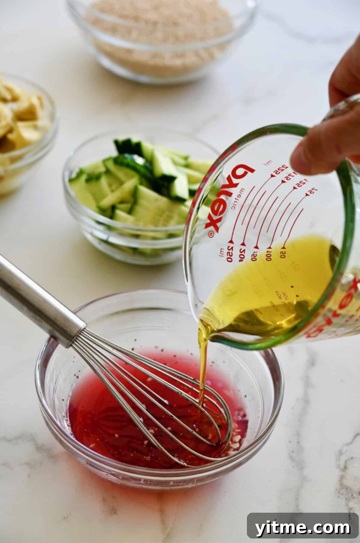 Pouring golden olive oil from a clear liquid measuring cup into a glass bowl, where it joins a mixture of red wine vinegar and lemon juice, beginning the emulsification process for a homemade red wine vinaigrette.