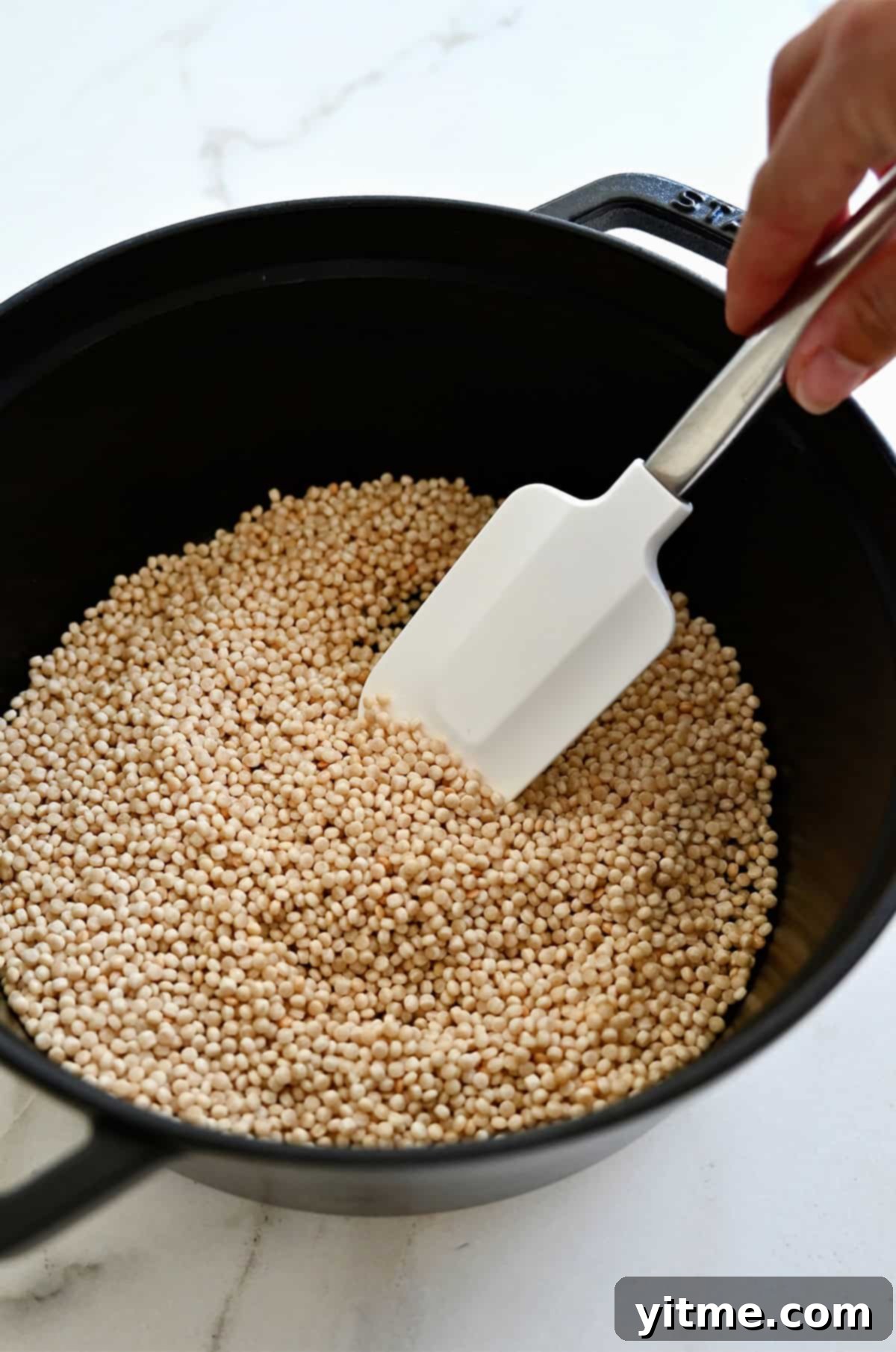Close-up shot of pearl couscous being toasted in a medium saucepan with a white spatula, turning lightly golden for enhanced nutty flavor.