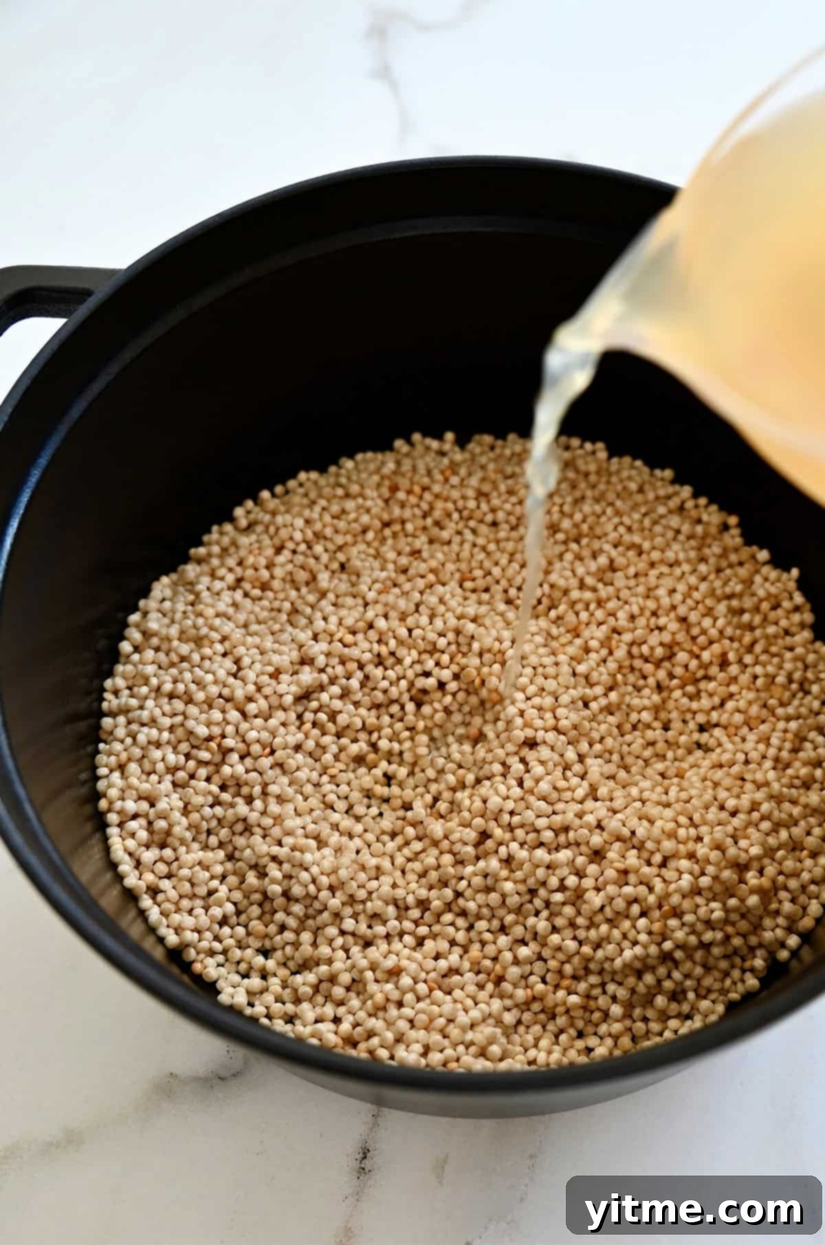 A stream of clear chicken broth being poured from a liquid measuring cup into a saucepot containing toasted pearl couscous, signifying the next step in cooking the couscous.