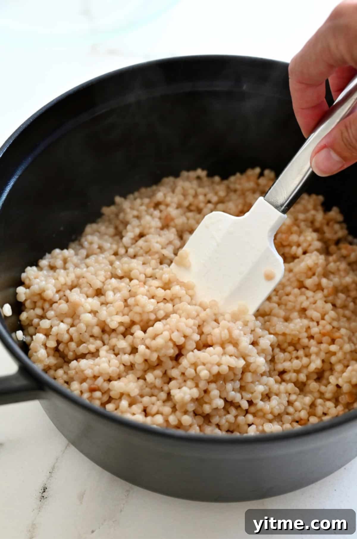 Cooked pearl couscous in a saucepot, fluffed gently with a white spatula, appearing perfectly tender and ready for the next step in the salad preparation.