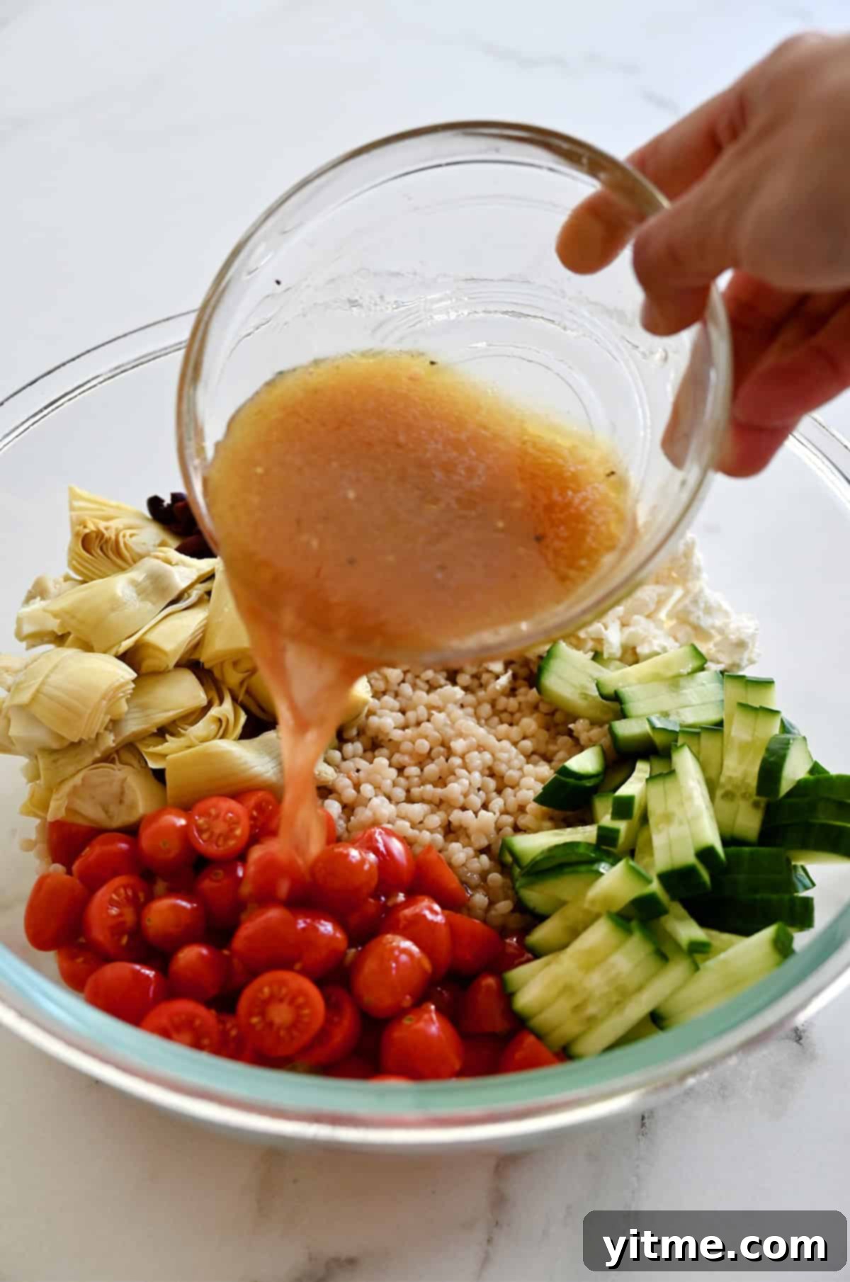 A small bowl of vibrant red wine vinaigrette being poured over a colorful Mediterranean couscous salad in a large bowl, featuring pearl couscous, halved cherry tomatoes, sliced cucumbers, chopped artichoke hearts, sliced Kalamata olives, and crumbled feta.