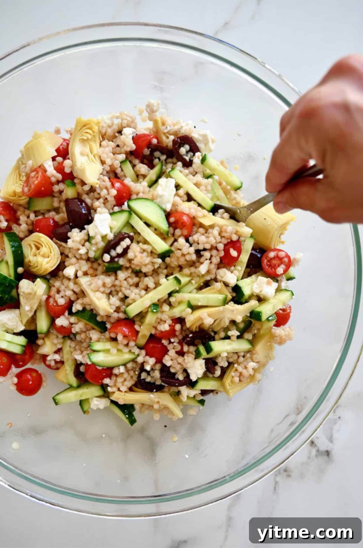 A pair of serving spoons actively tossing a Mediterranean couscous salad with red wine vinaigrette in a large white bowl, mixing all the colorful ingredients thoroughly.