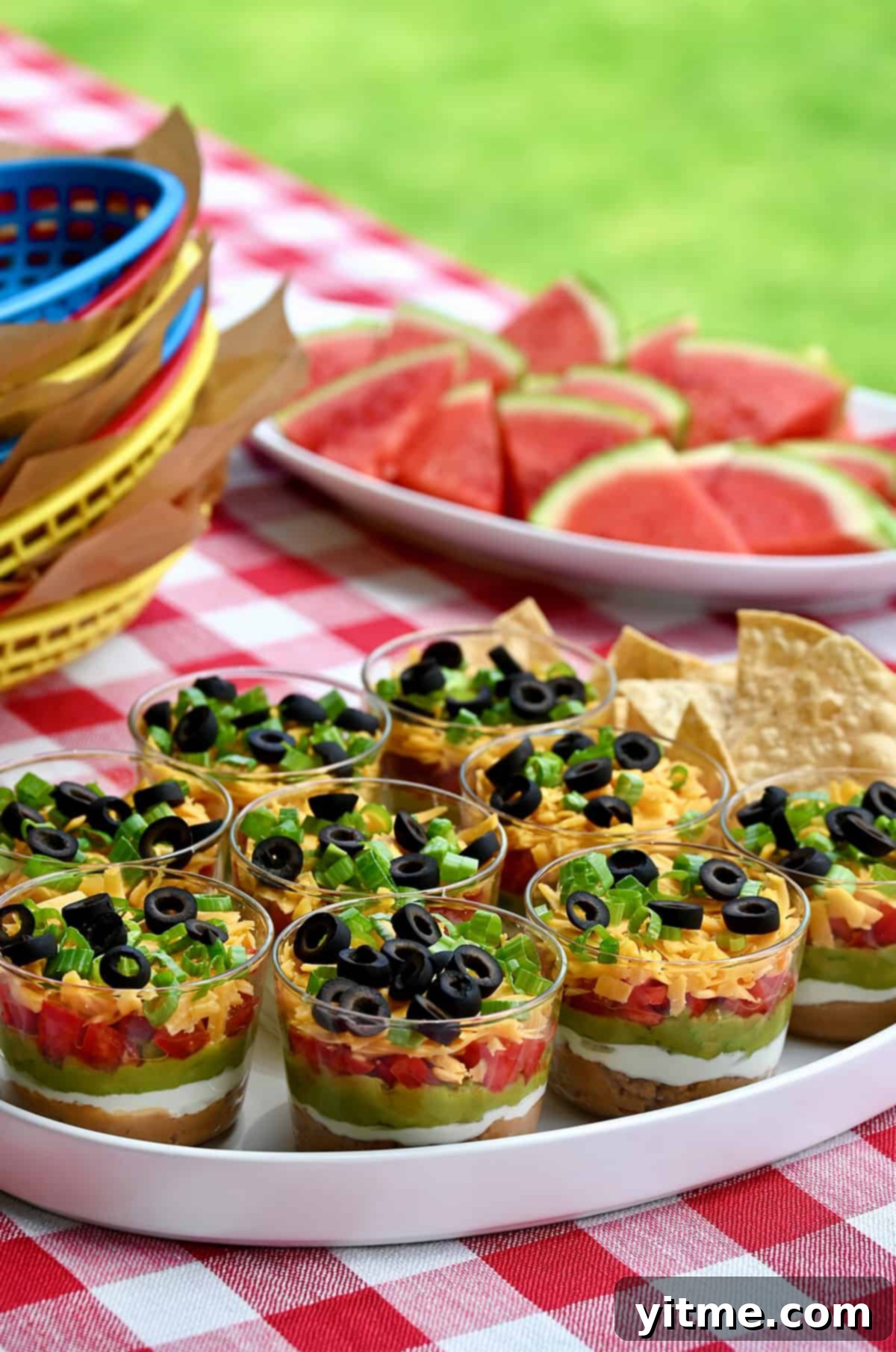 Individual 7-layer dips served in clear mini cups on a red and white checkered tablecloth, perfect for summer parties or BBQs.