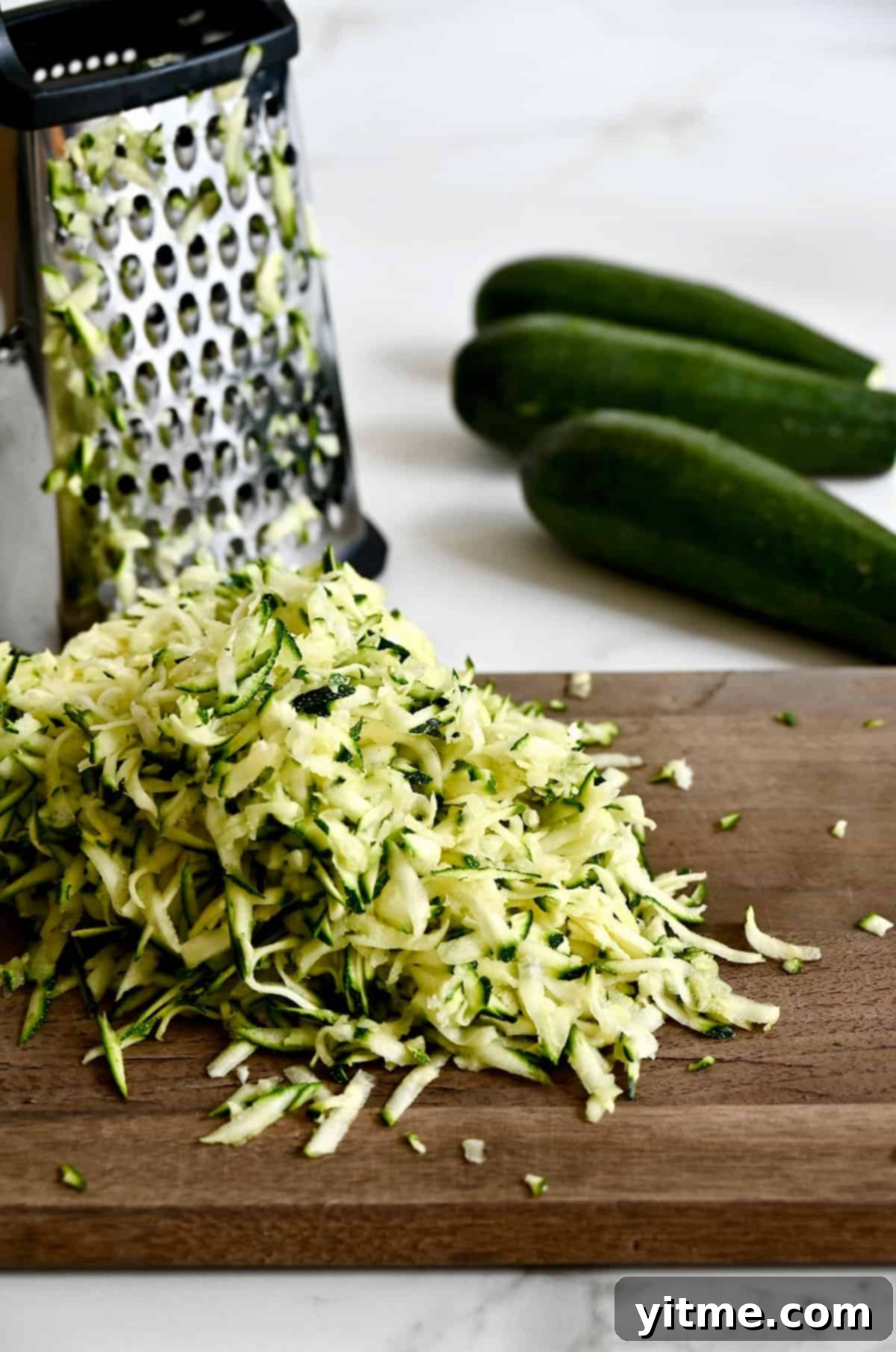 Shredded zucchini on a cutting board next to a box grater.