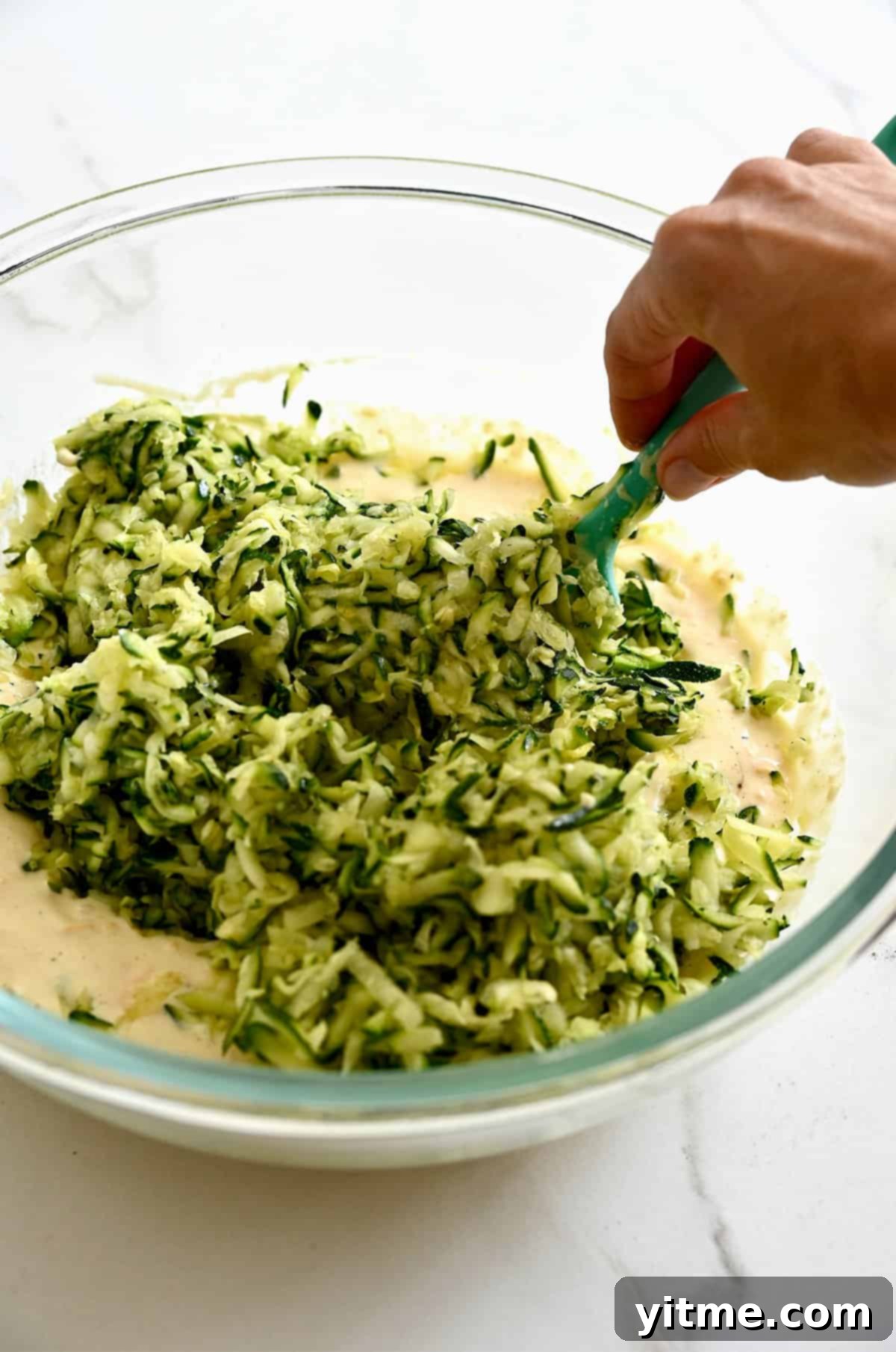 Grated zucchini being stirred into the cheesy mixture with a spatula.