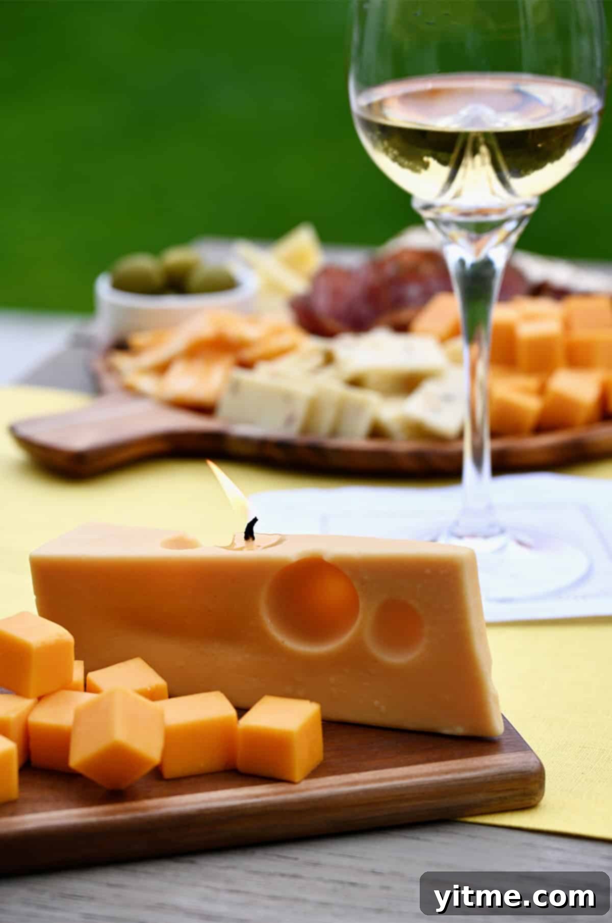 Cheese candle on a charcuterie board with cubed cheddar cheese and a glass of white wine nearby, adding a whimsical touch to the spread.