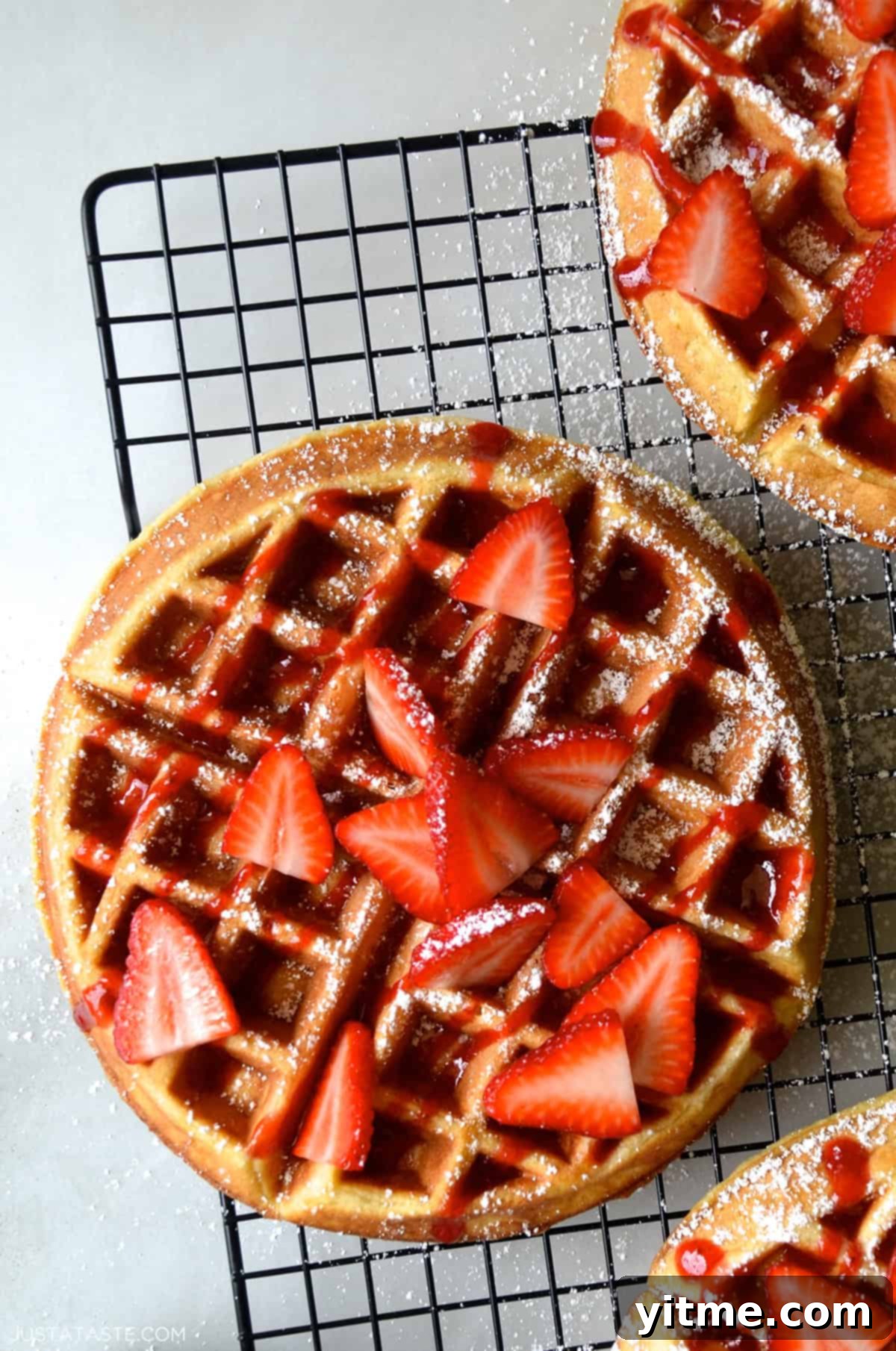 A homemade buttermilk waffle topped with golden maple syrup, fresh strawberries, and a light dusting of powdered sugar, presented on a wire cooling rack.