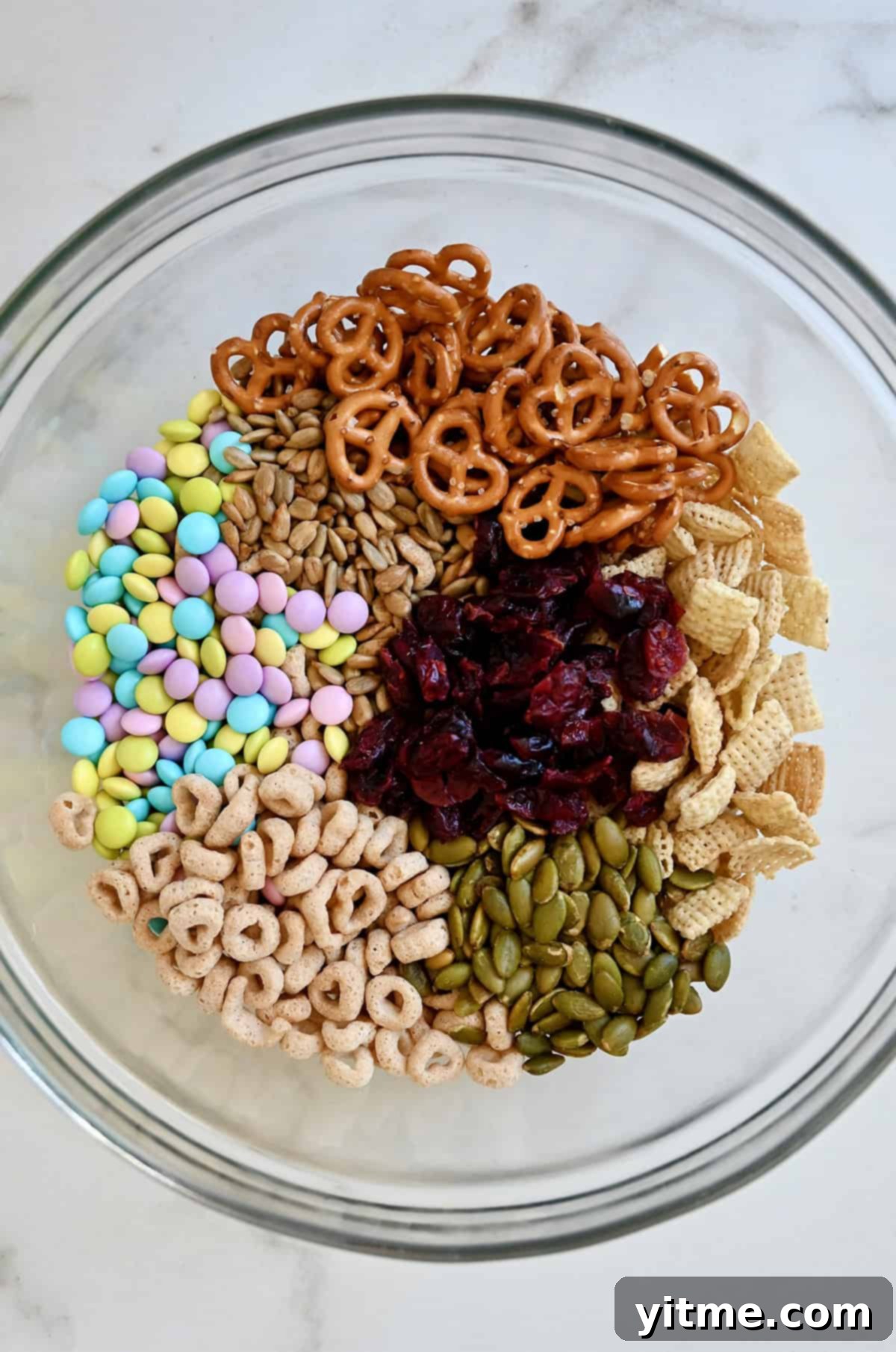 Pastel colored M&Ms, sunflower seeds, mini pretzels, Chex cereal, pumpkin seeds, Cheerios and dried cranberries in a bowl.