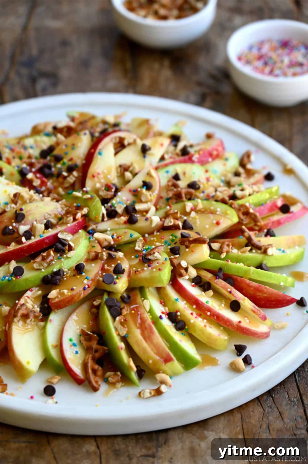 Caramel apple nachos starring thin slices of apples topped with a thick drizzle of caramel sauce, mini chocolate chips, chopped peanuts, chopped pretzels and rainbow sprinkles.