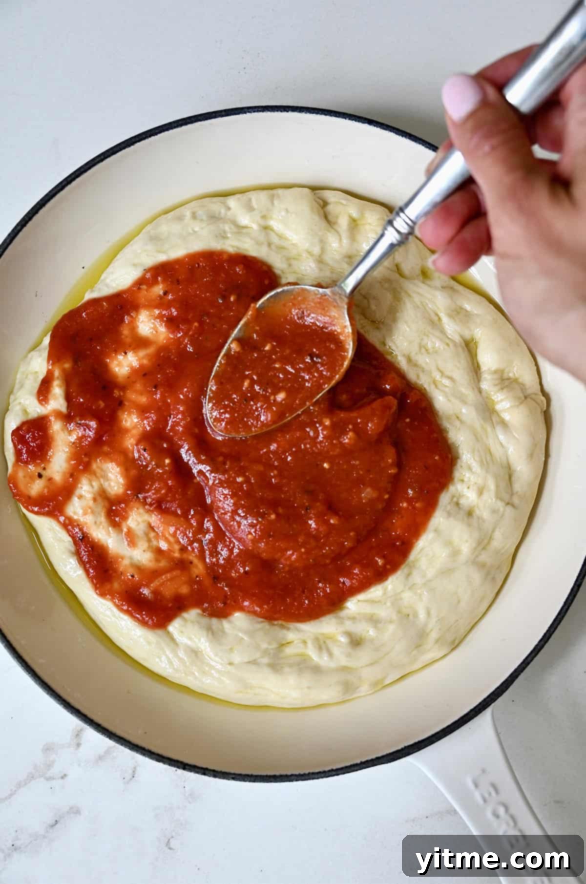 Spreading homemade marinara sauce evenly across the pizza dough in a cast iron pan, extending all the way to the edges.