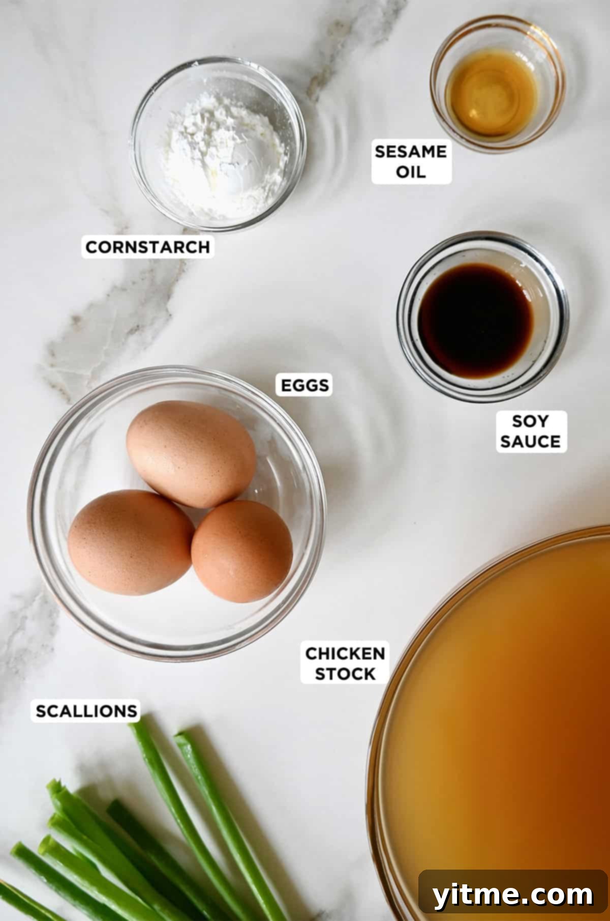 Fresh ingredients laid out for making egg drop soup, including a container of chicken stock, a bowl of beaten eggs, a bottle of soy sauce, sesame oil, cornstarch, and a bunch of scallions.