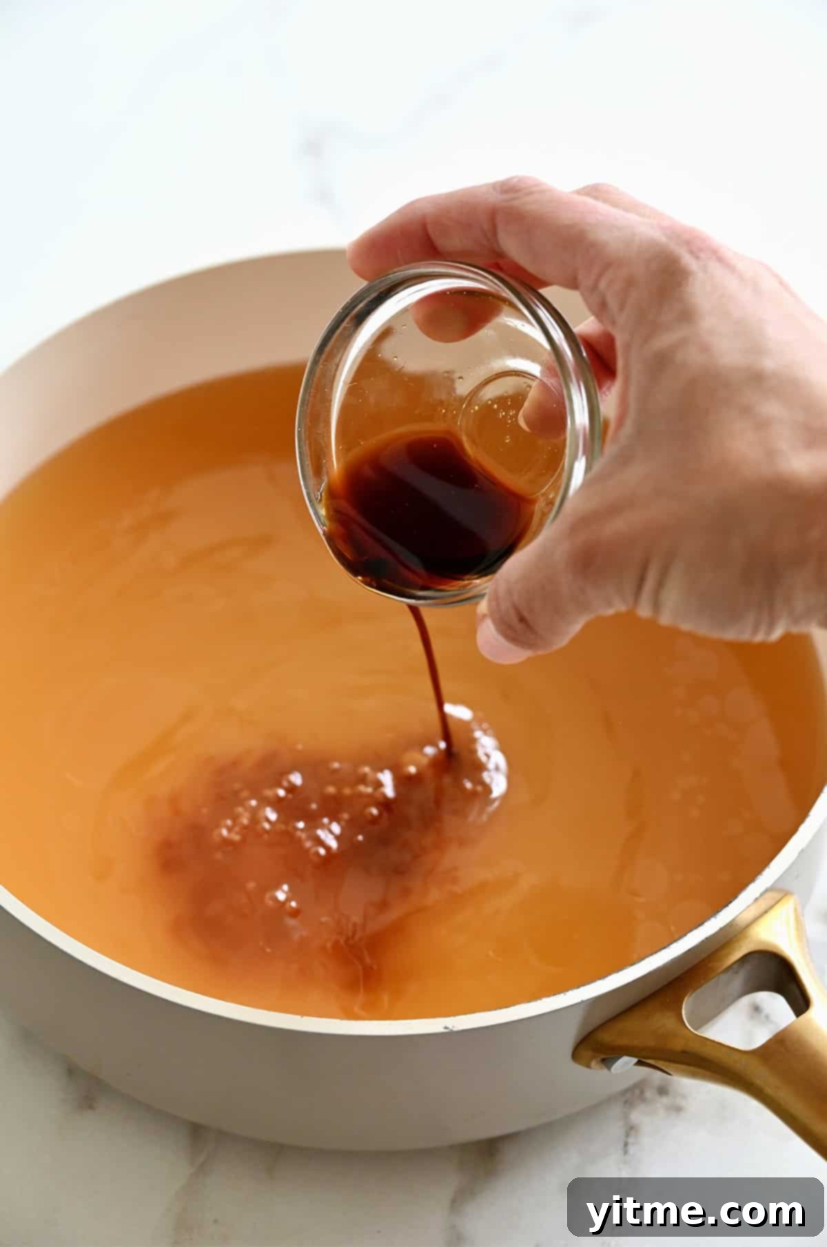 A small glass bowl containing soy sauce and sesame oil being poured into a large stock pot with chicken stock on a stove.