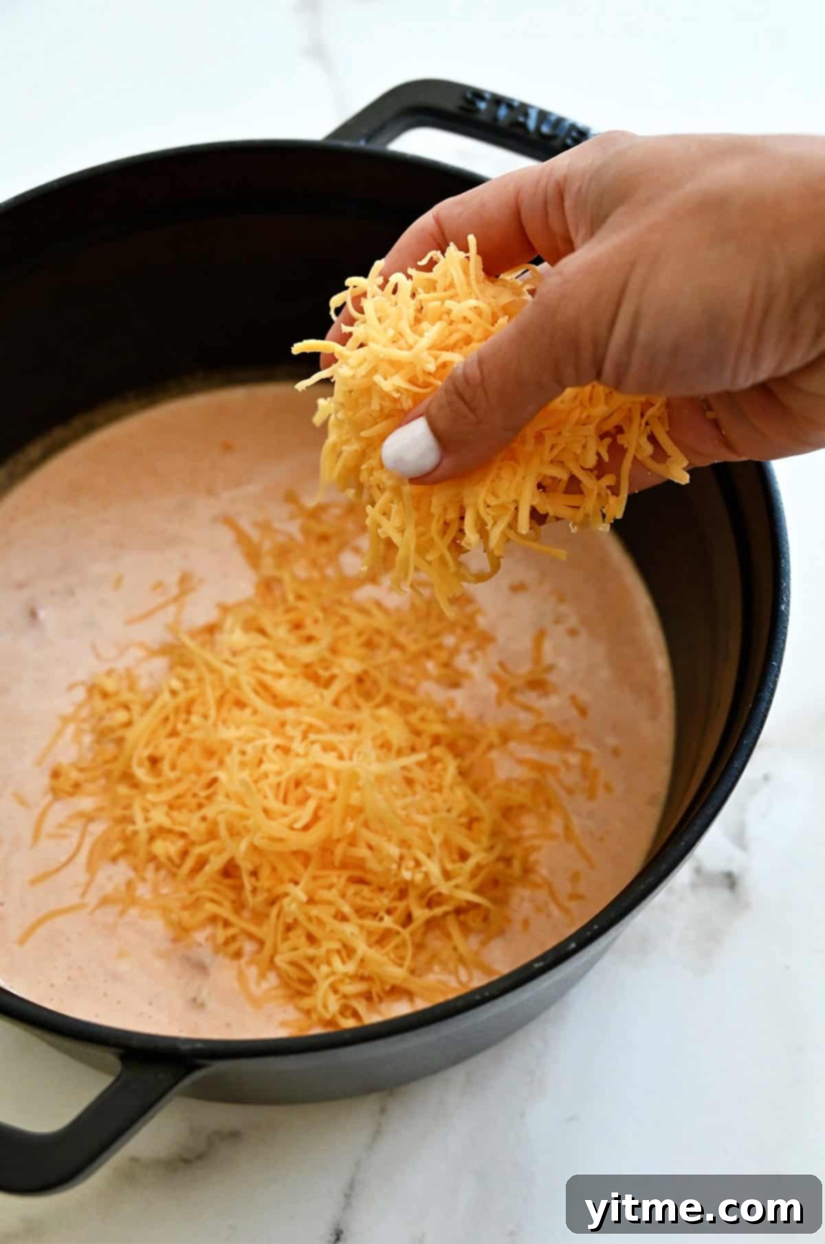 Shredded cheddar cheese is being added to a saucepan to make warm queso sauce.