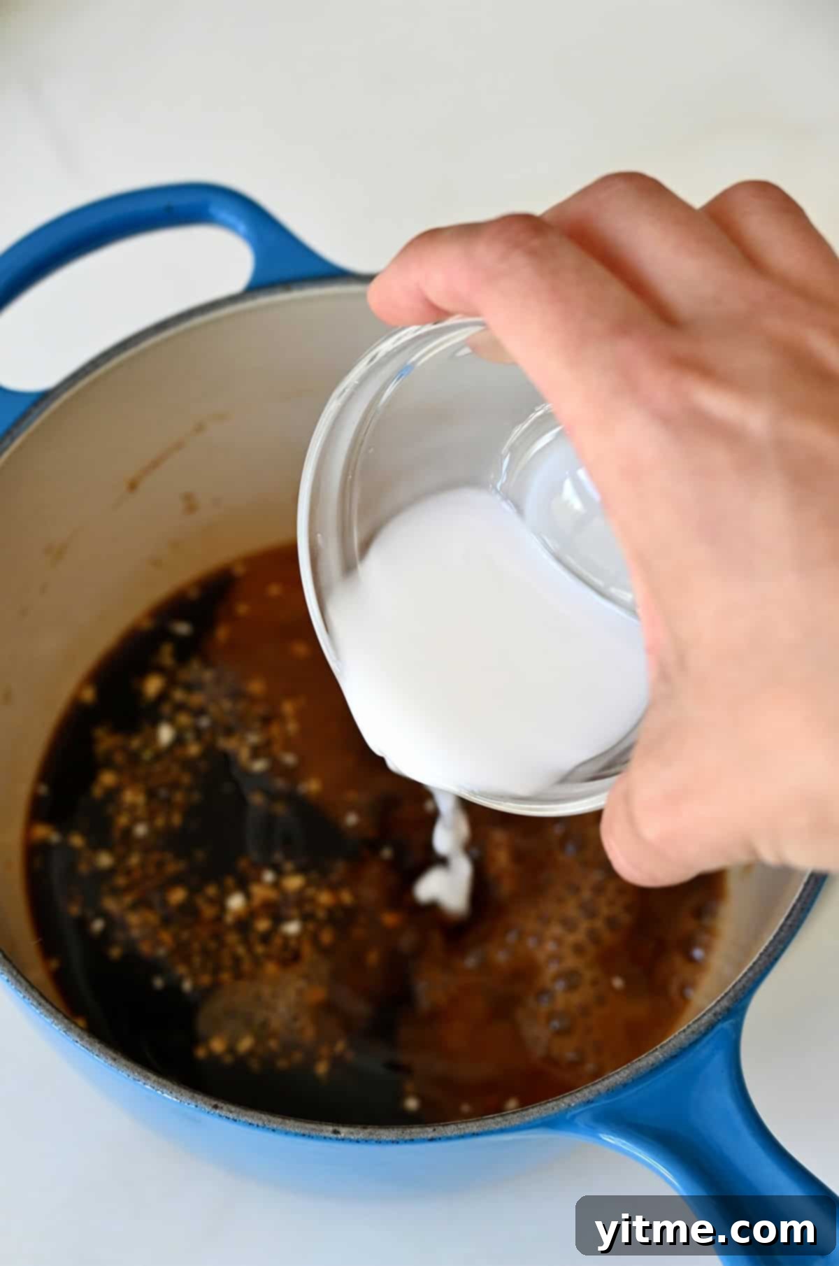 Adding a cornstarch slurry to a simmering homemade teriyaki sauce in a saucepan, demonstrating the thickening process.