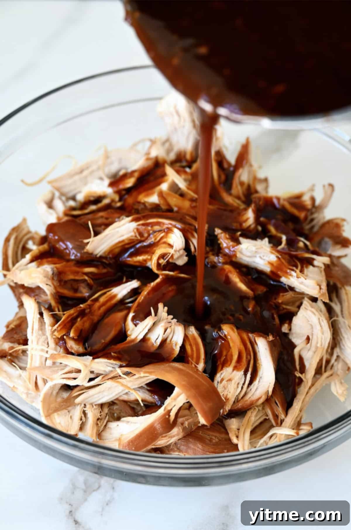 Homemade teriyaki sauce being poured generously over tender pulled chicken in a serving bowl, showcasing its rich, glossy texture.