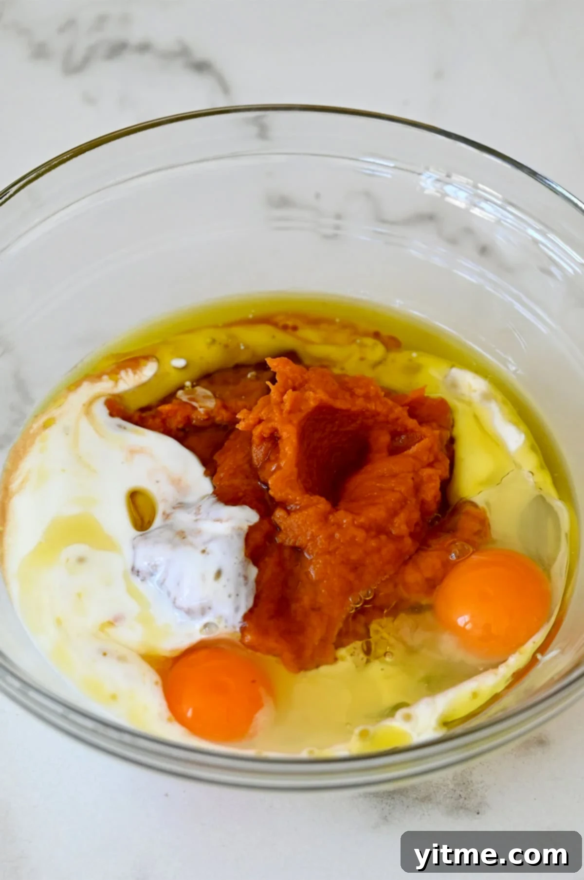 Wholesome Pumpkin Delights 6 A glass bowl filled with the wet ingredients essential for creating moist and flavorful pumpkin muffins. The mix consists of pumpkin purée, eggs, buttermilk, vanilla extract, and olive oil, thoroughly whisked together.