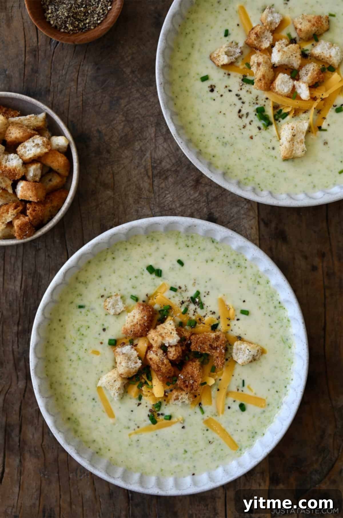 Creamy Broccoli Cheddar Soup 12 Two bowls containing creamy homemade broccoli cheddar soup topped with crunchy croutons, shredded cheddar cheese and chopped fresh chives.