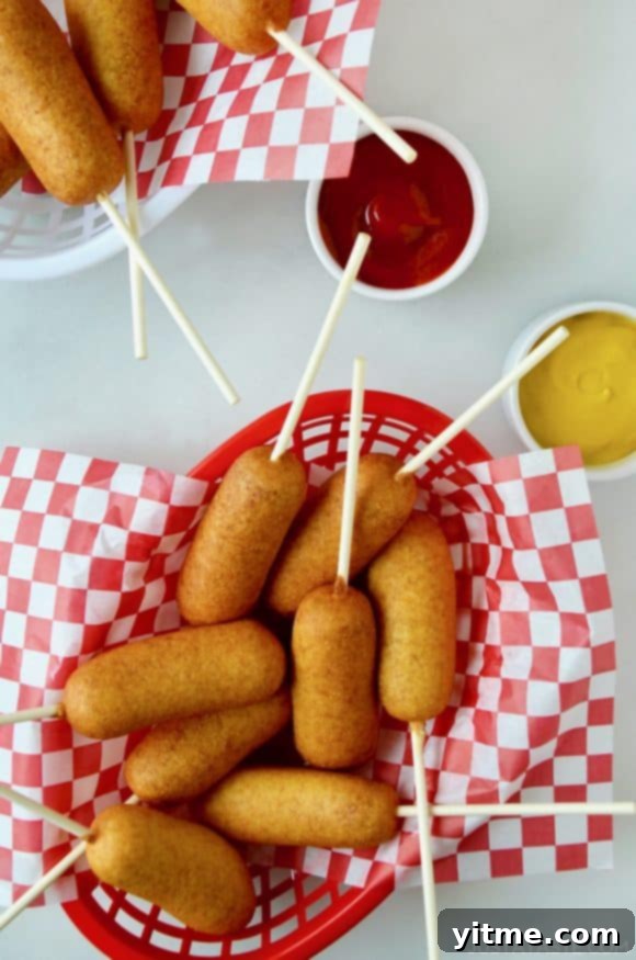 A red food basket filled with Easy Homemade Mini Corn Dogs, served alongside small ramekins of ketchup and mustard for dipping.