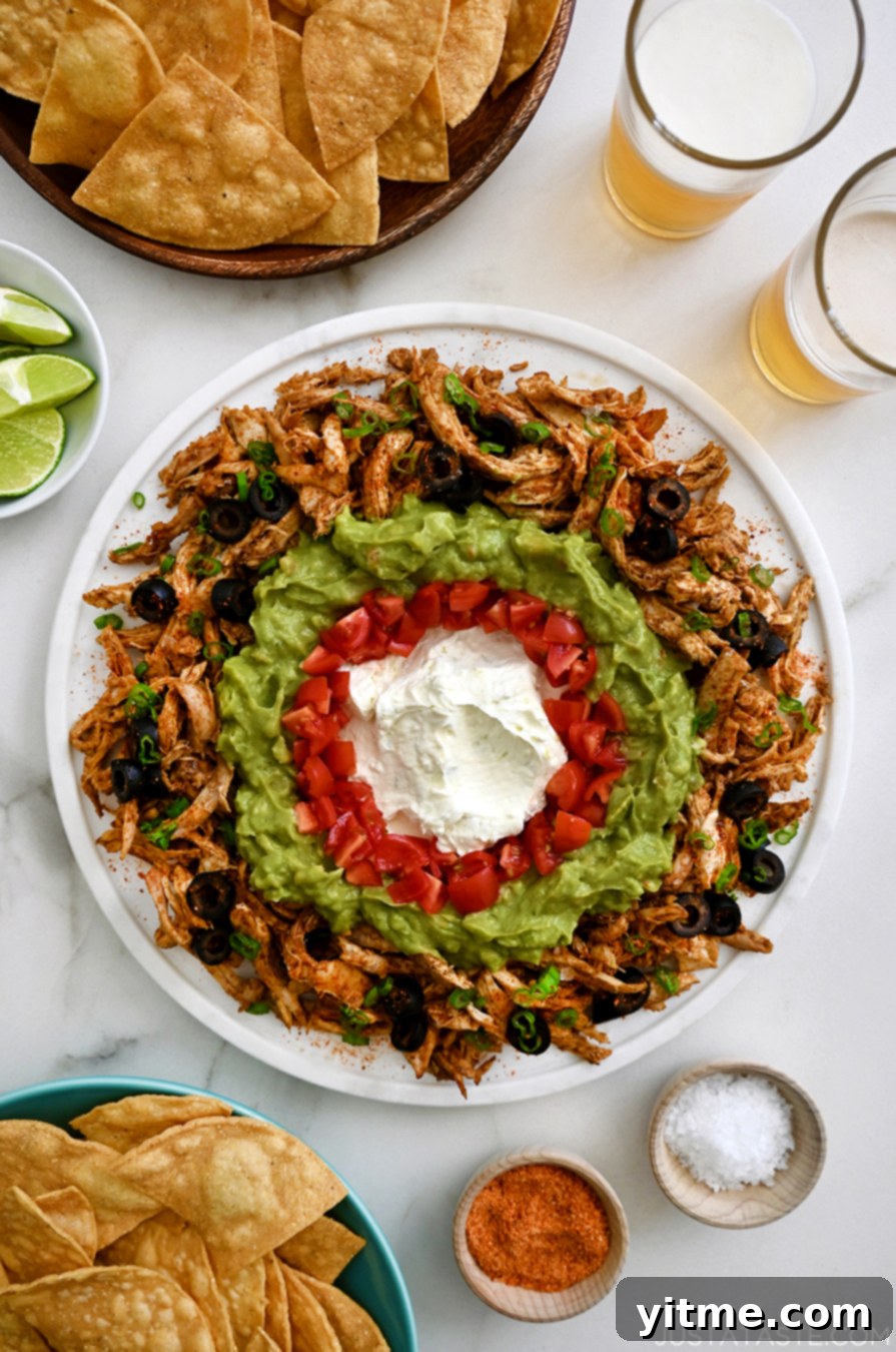 A top-down view of a white platter featuring Rotisserie Chicken Taco Dip, surrounded by fresh limes and bowls of tortilla chips, perfect for dipping.
