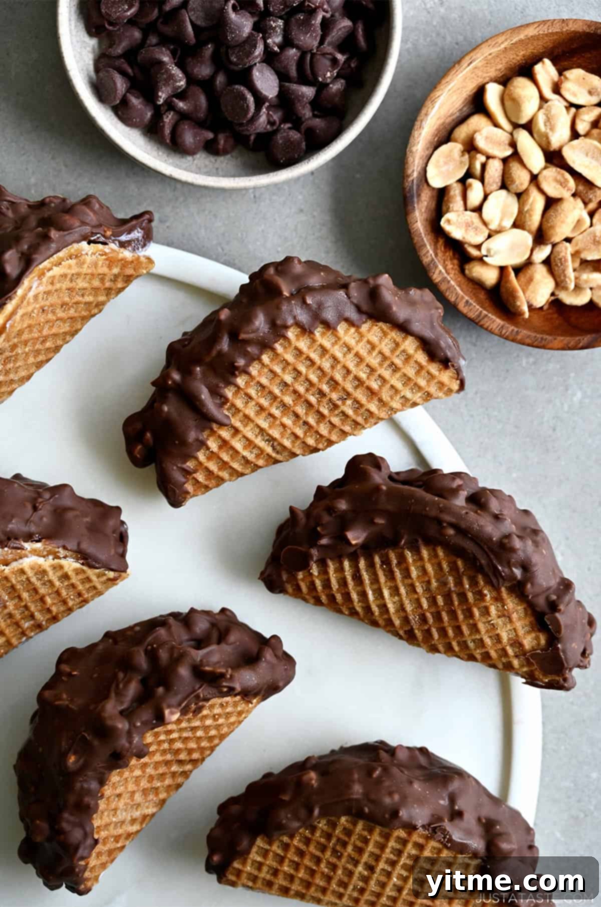 Homemade choco tacos on a round serving tray, next to a bowl of chocolate chips and another bowl of peanuts for topping.