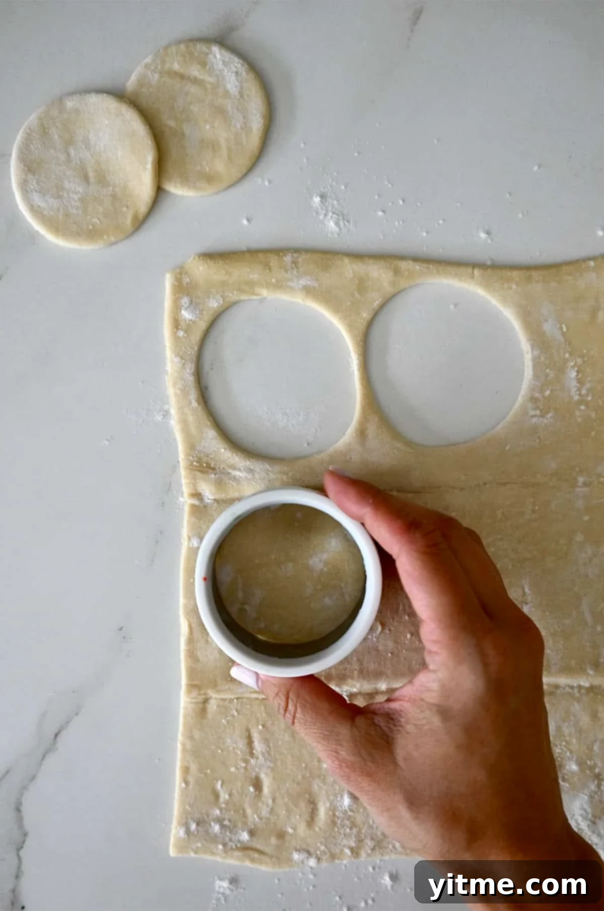 A hand holding a circular cookie cutter cuts out puff pastry circles.