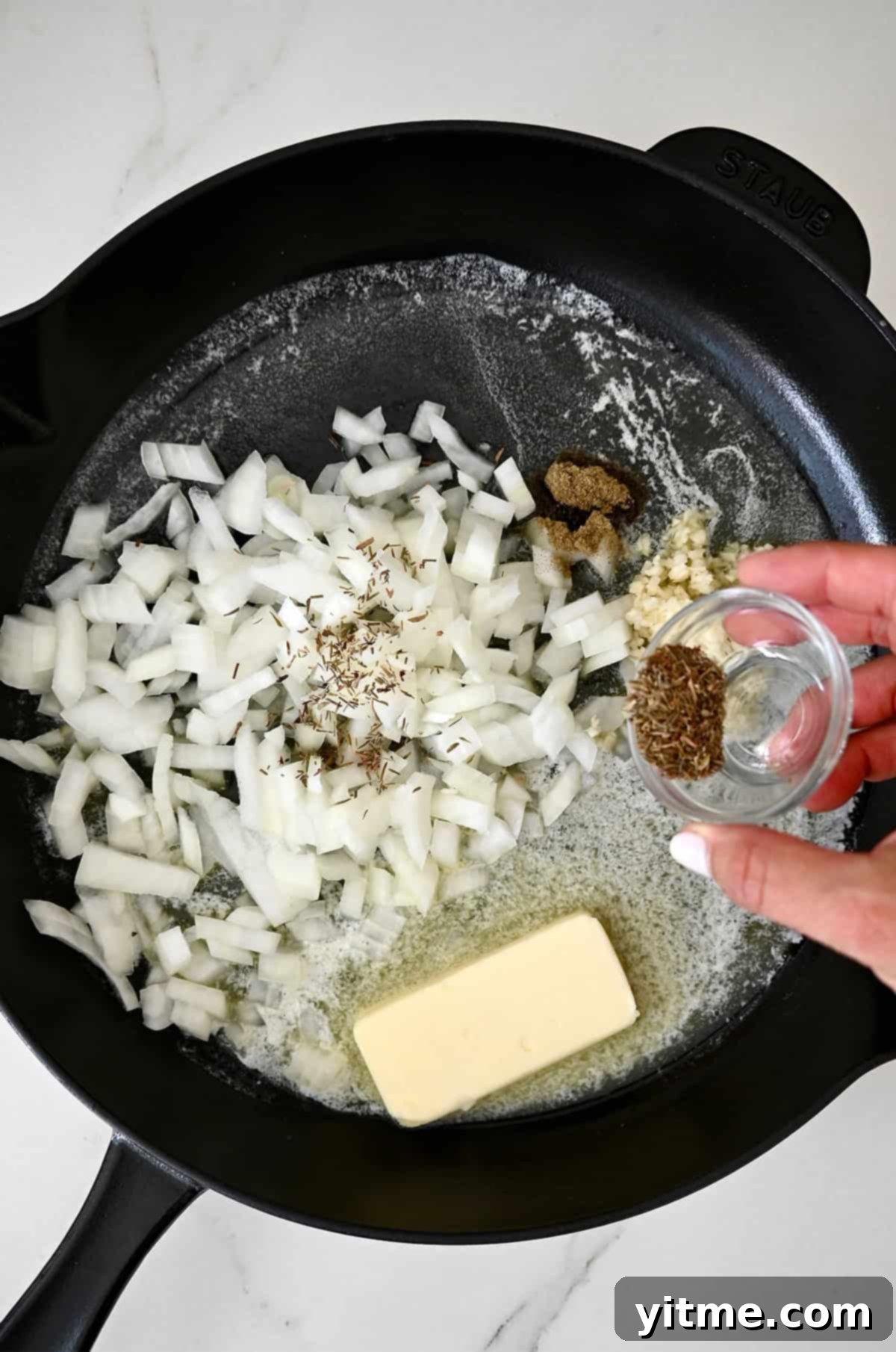 Dried herbs are being added to a cast-iron skillet with diced onions, garlic, and half a stick of butter.