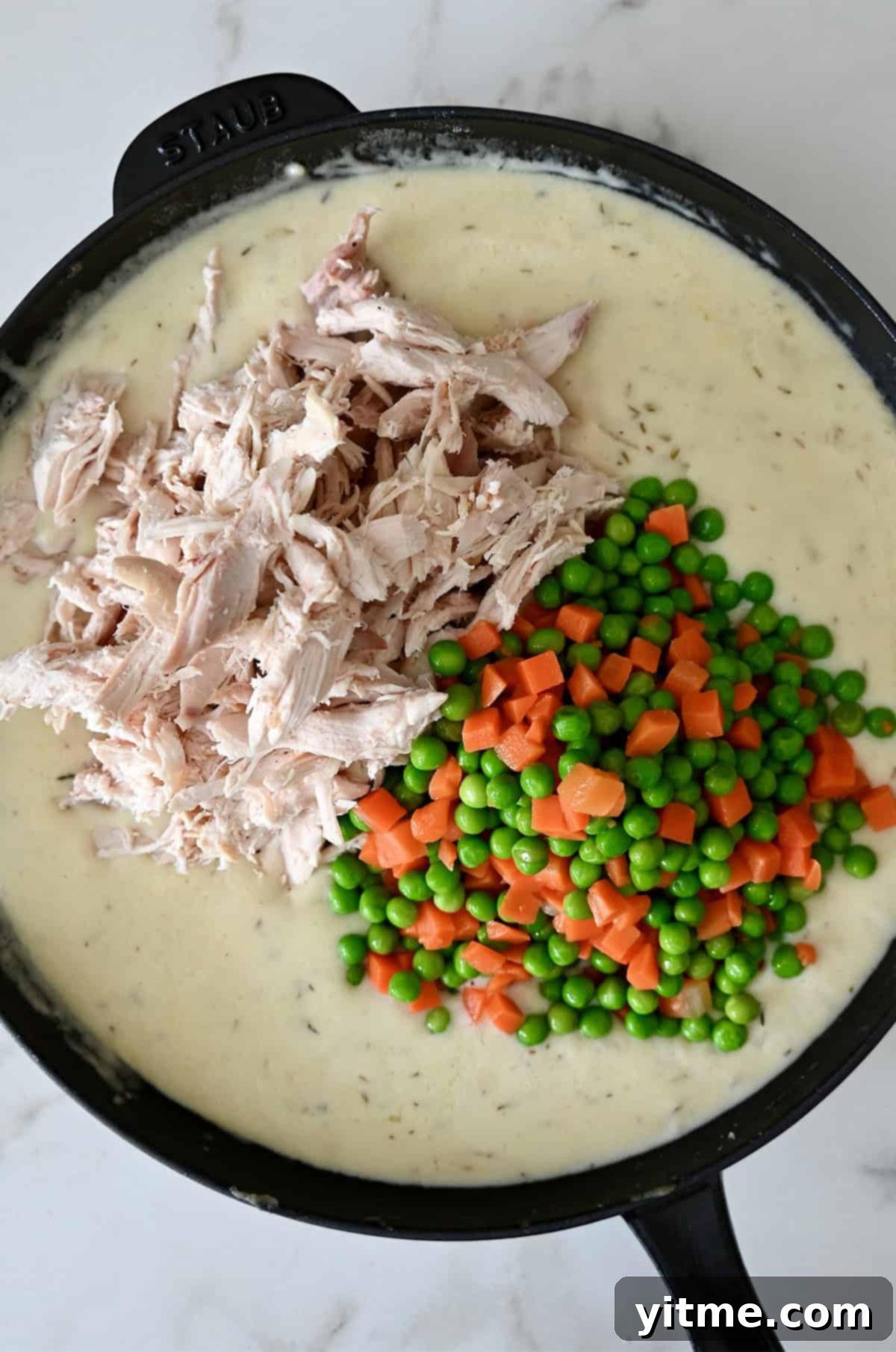 Shredded rotisserie chicken, peas and carrots being folded into creamy sauce in a skillet.