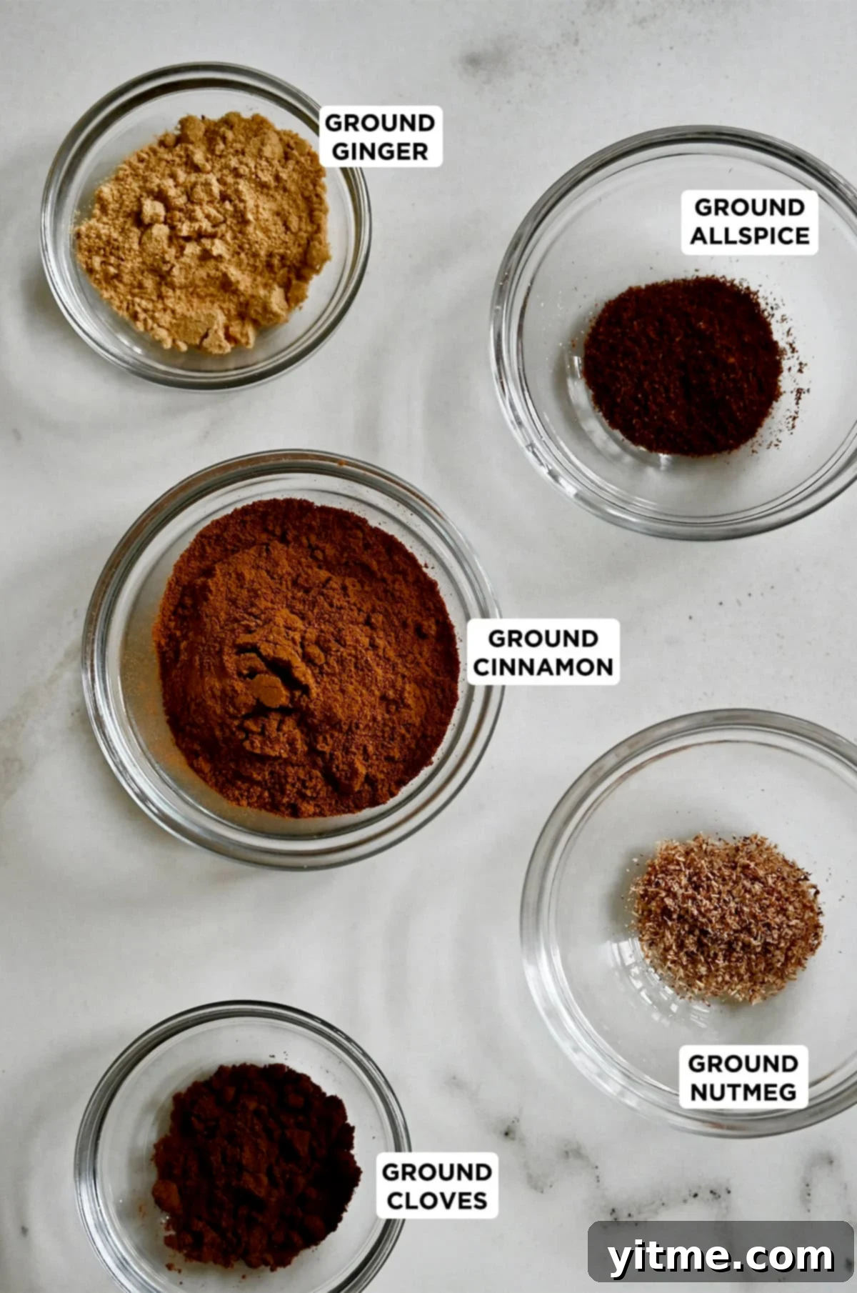 Five small glass bowls, each holding a distinct ground spice – cinnamon, ginger, nutmeg, cloves, and allspice – the key components for a homemade pumpkin pie spice blend.