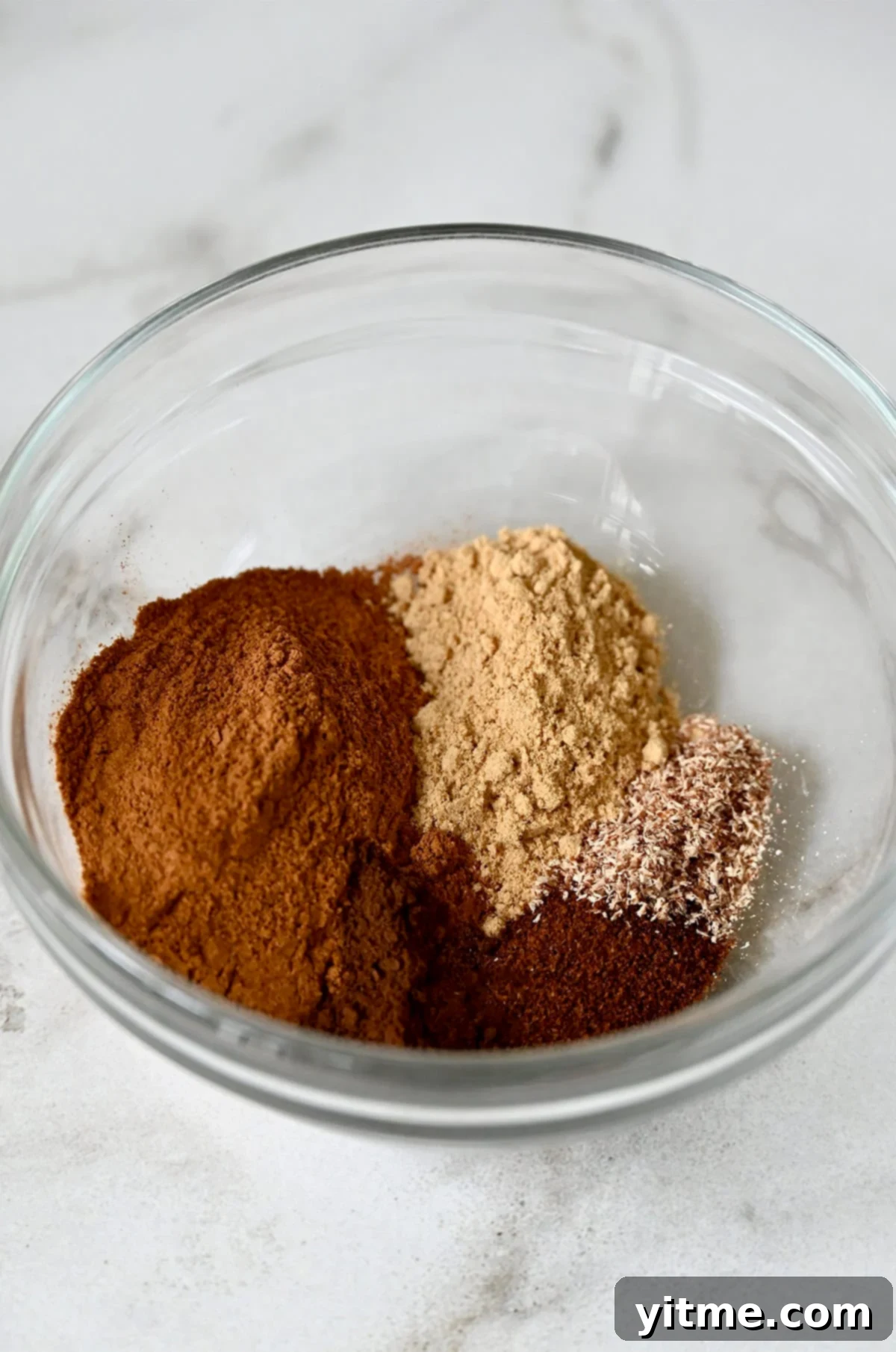A vibrant mix of ground cinnamon, allspice, ginger, nutmeg, and cloves fills a small clear glass bowl, showcasing the rich colors of the freshly combined pumpkin pie spice.
