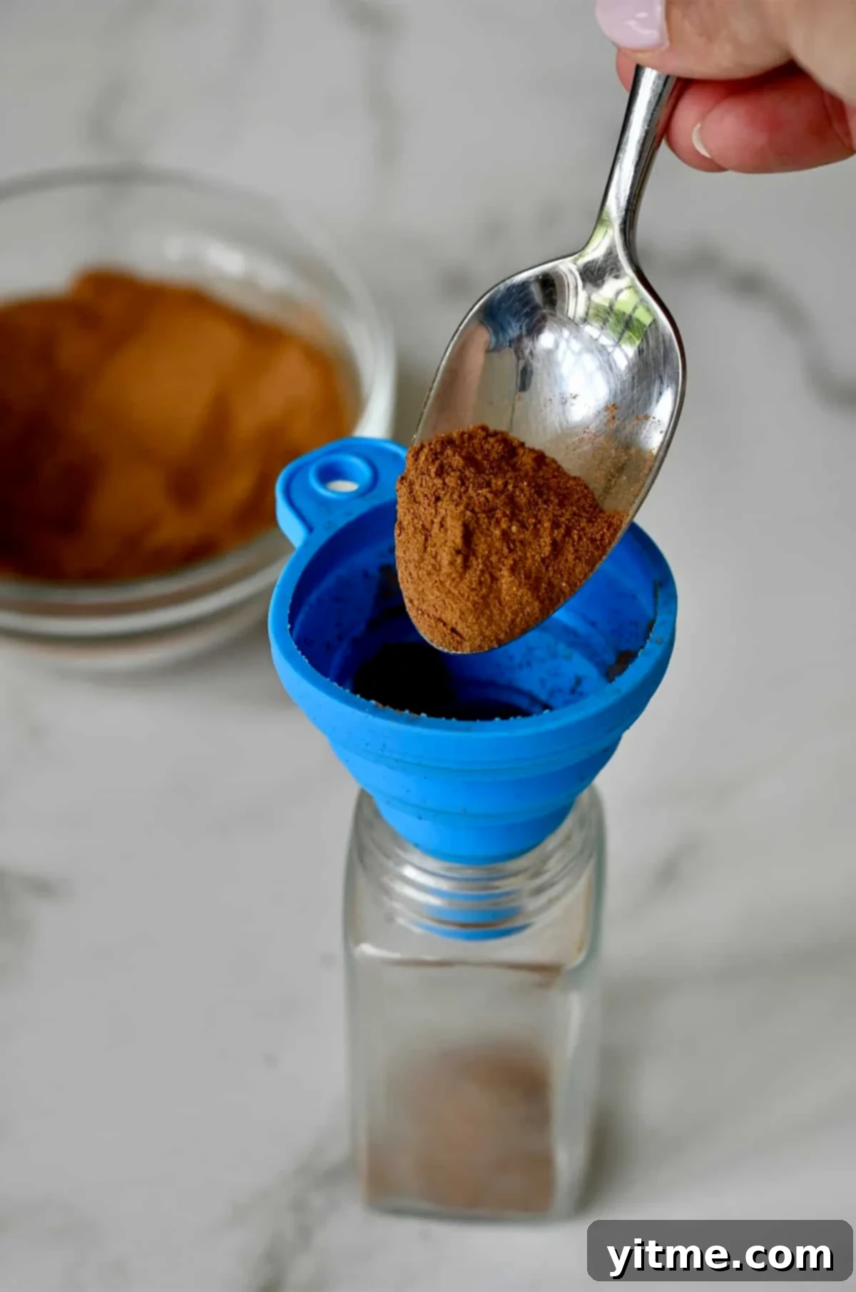 A hand carefully spoons homemade pumpkin pie spice into a small glass jar using a funnel, ensuring no precious spice is wasted.