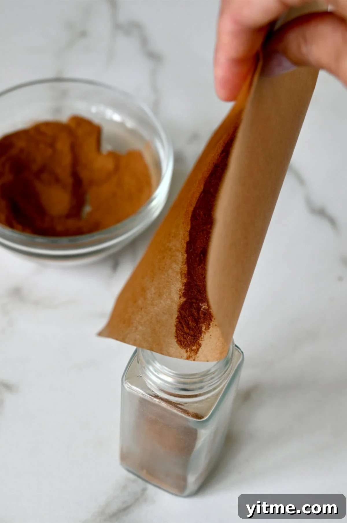 A simple yet effective technique: using a piece of folded parchment paper as a makeshift funnel to guide homemade pumpkin pie spice into a glass jar.