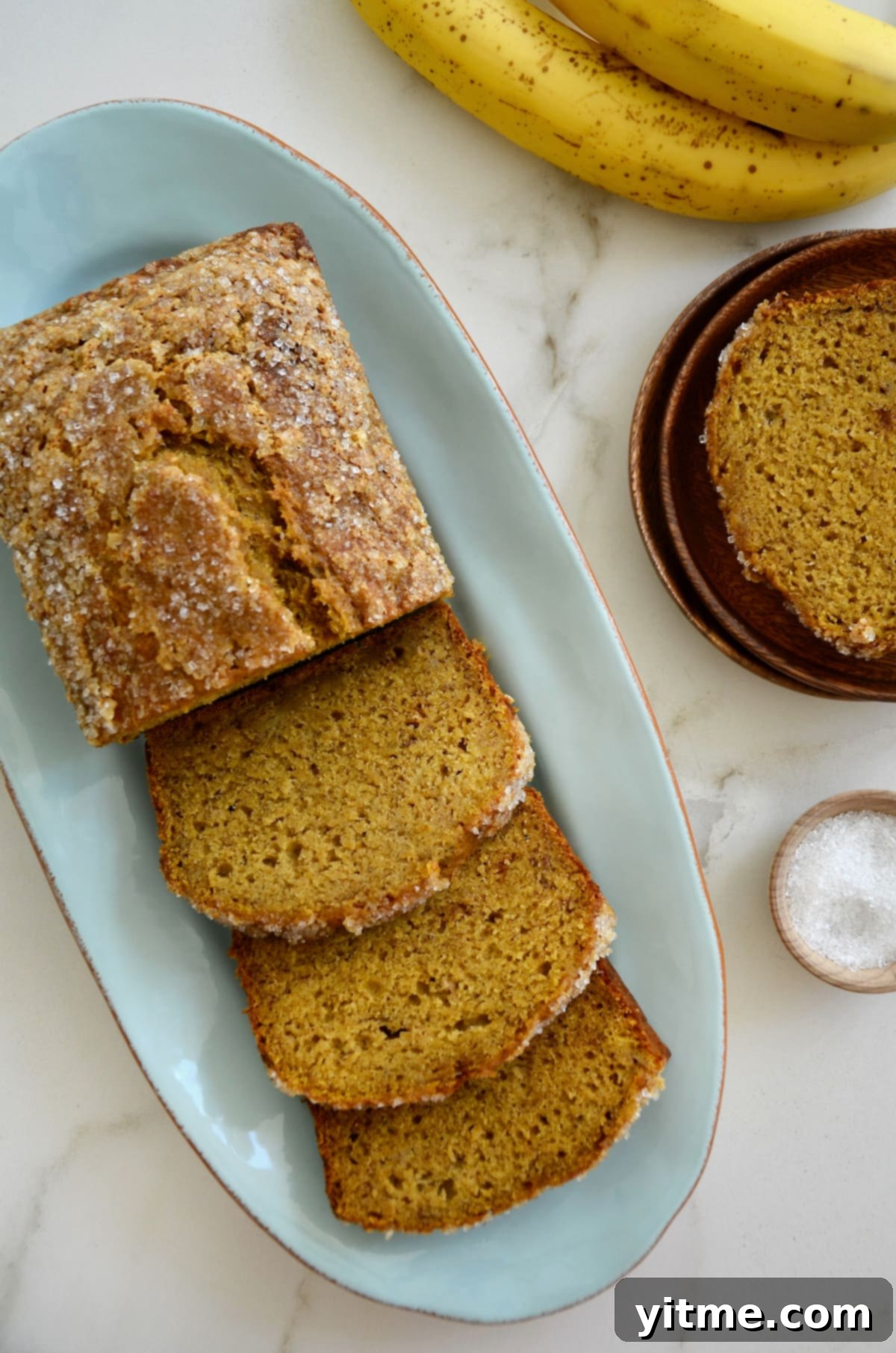 A beautifully baked loaf of Pumpkin Banana Bread sits on a blue platter, flanked by speckled brown bananas and a small dish of sanding sugar, inviting a taste of autumn.