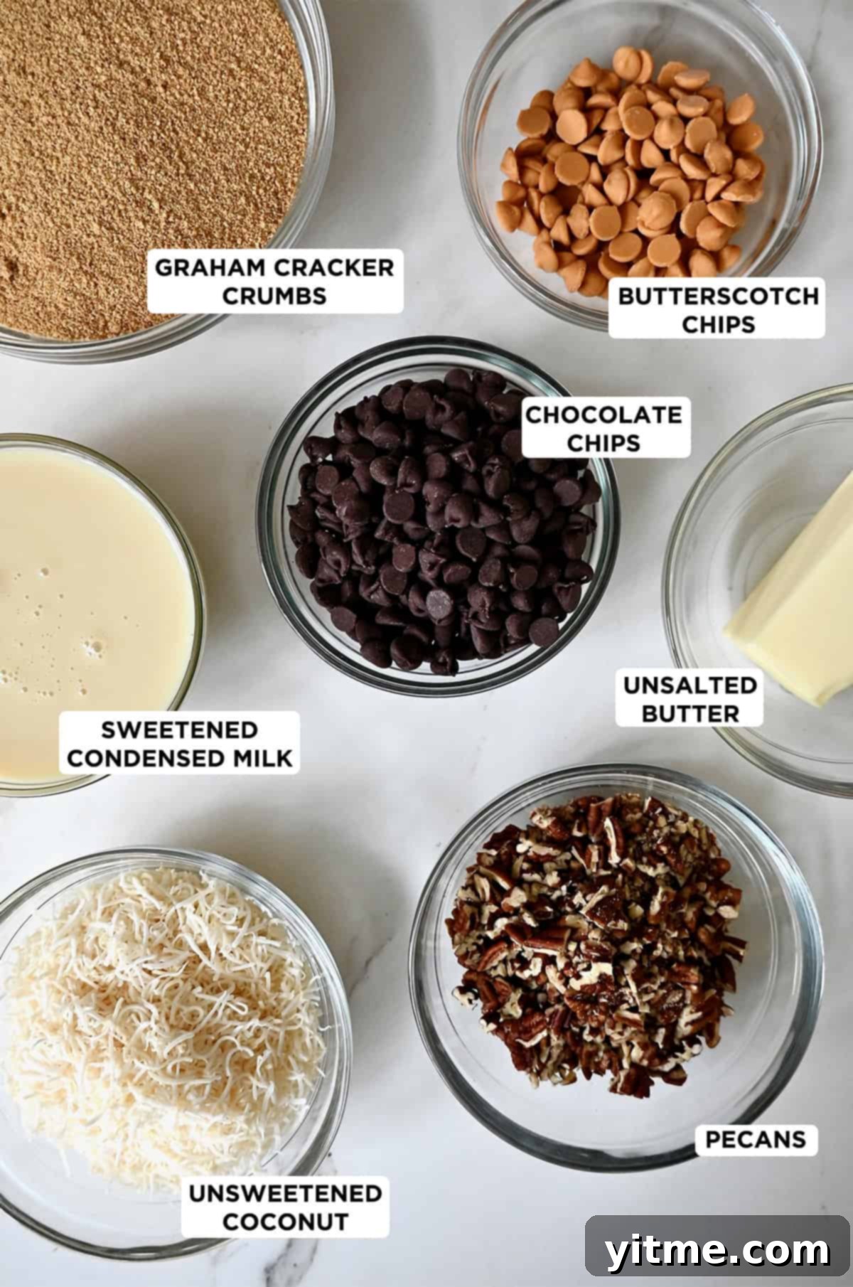Everything Cookie Bars 4 Glass bowls containing ingredients to make magic cookie bars, including crushed graham crackers, melted butter, butterscotch chips, sweetened condensed milk, flaked coconut, chopped pecans and semisweet chocolate chips.