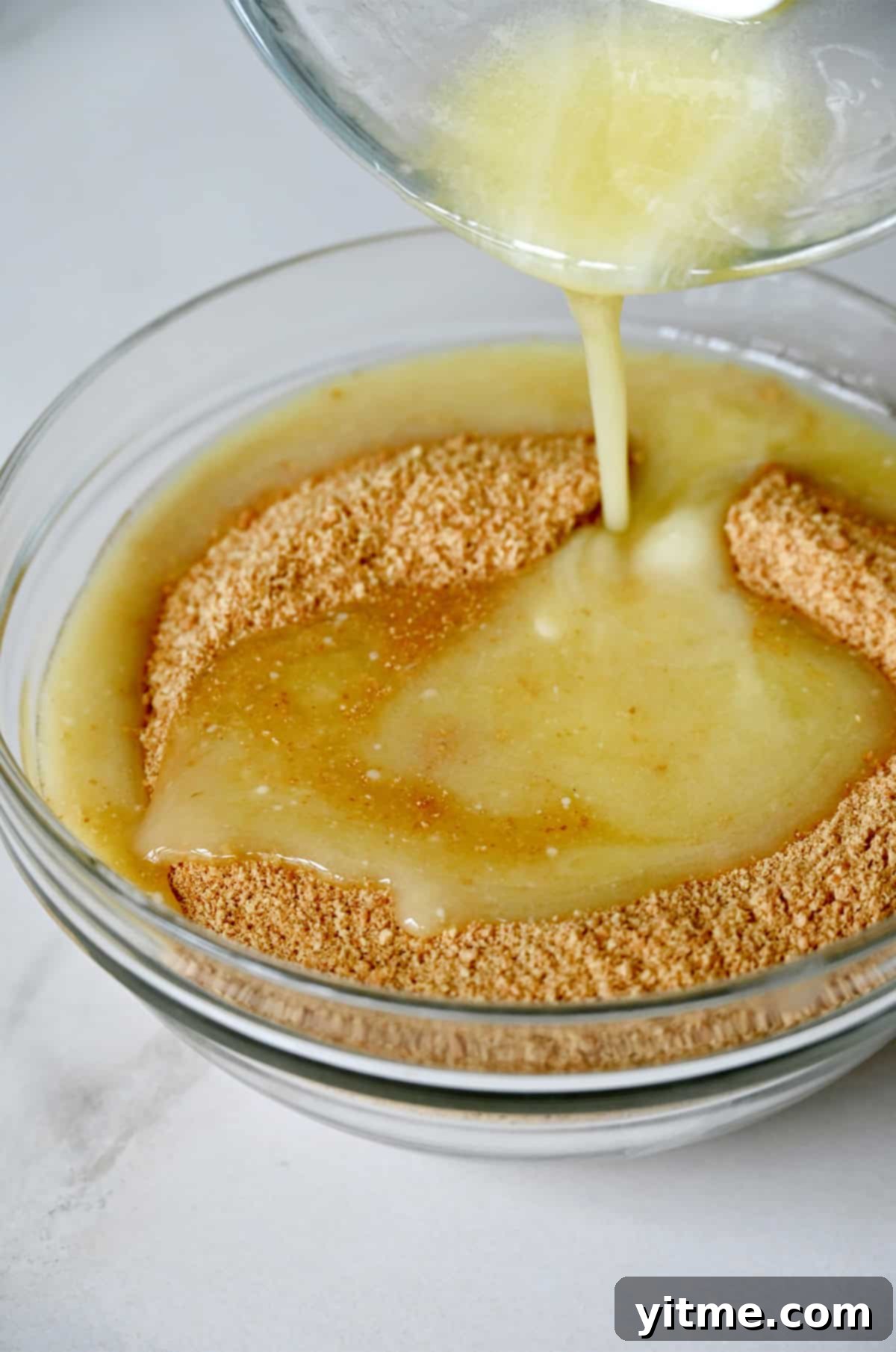 Everything Cookie Bars 6 Melted butter being poured into a bowl with crushed graham crackers, the first step for the crust.