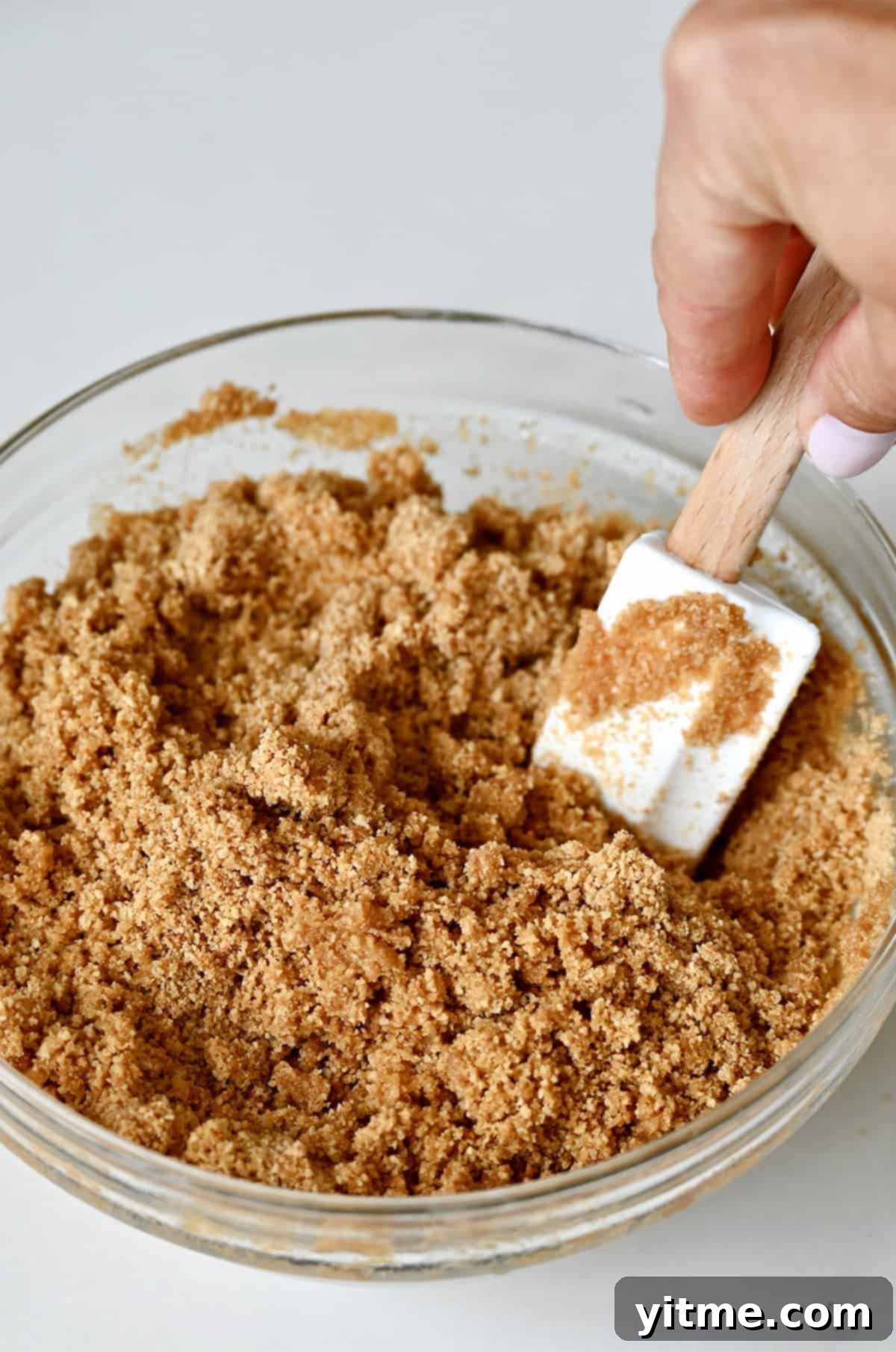 Everything Cookie Bars 7 Graham crackers and melted butter being mixed together with a spatula in a glass bowl, ensuring an even consistency.