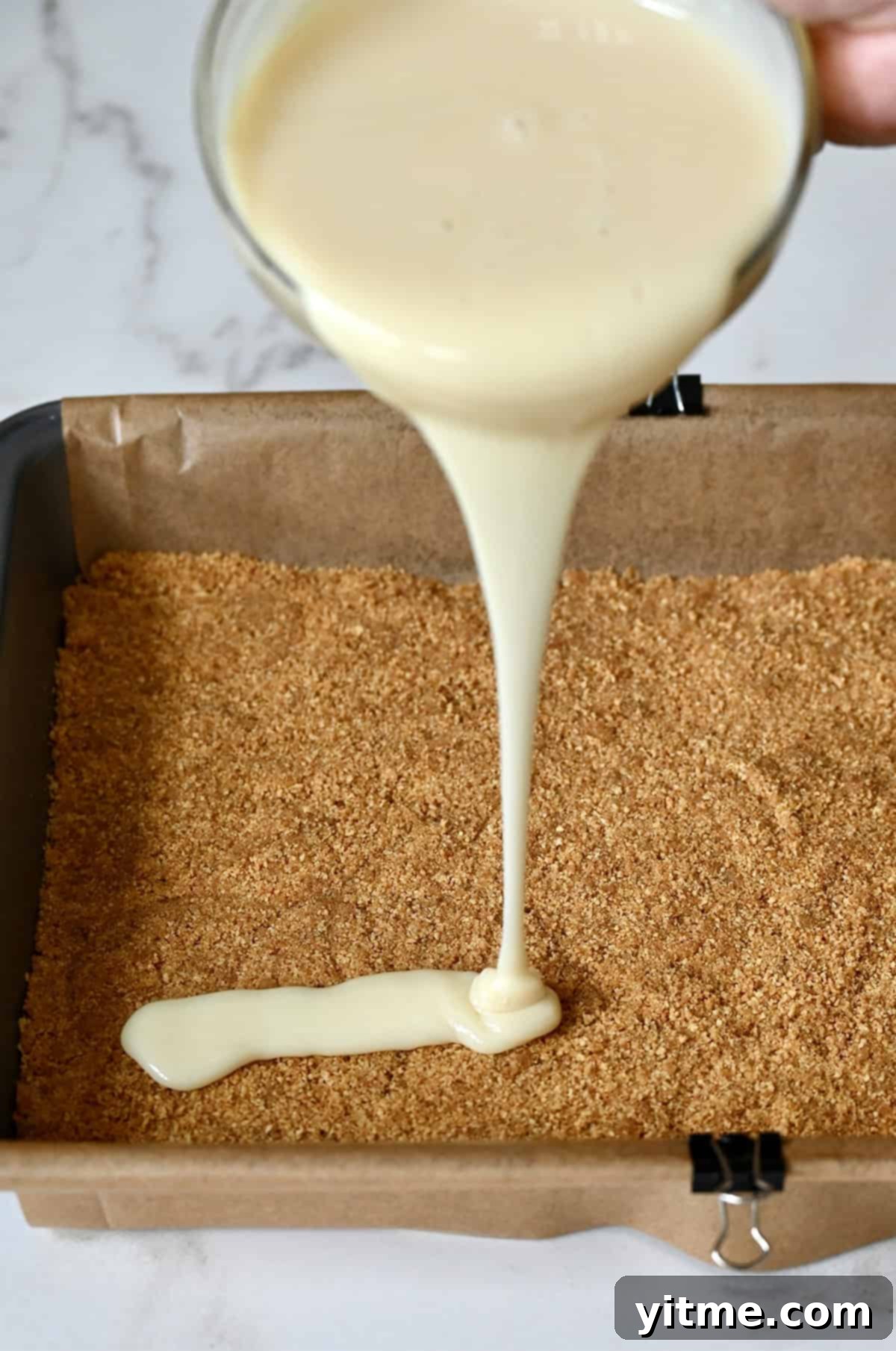 Everything Cookie Bars 9 Pouring warmed sweetened condensed milk atop a graham cracker crust in a baking pan, creating the sticky foundation.