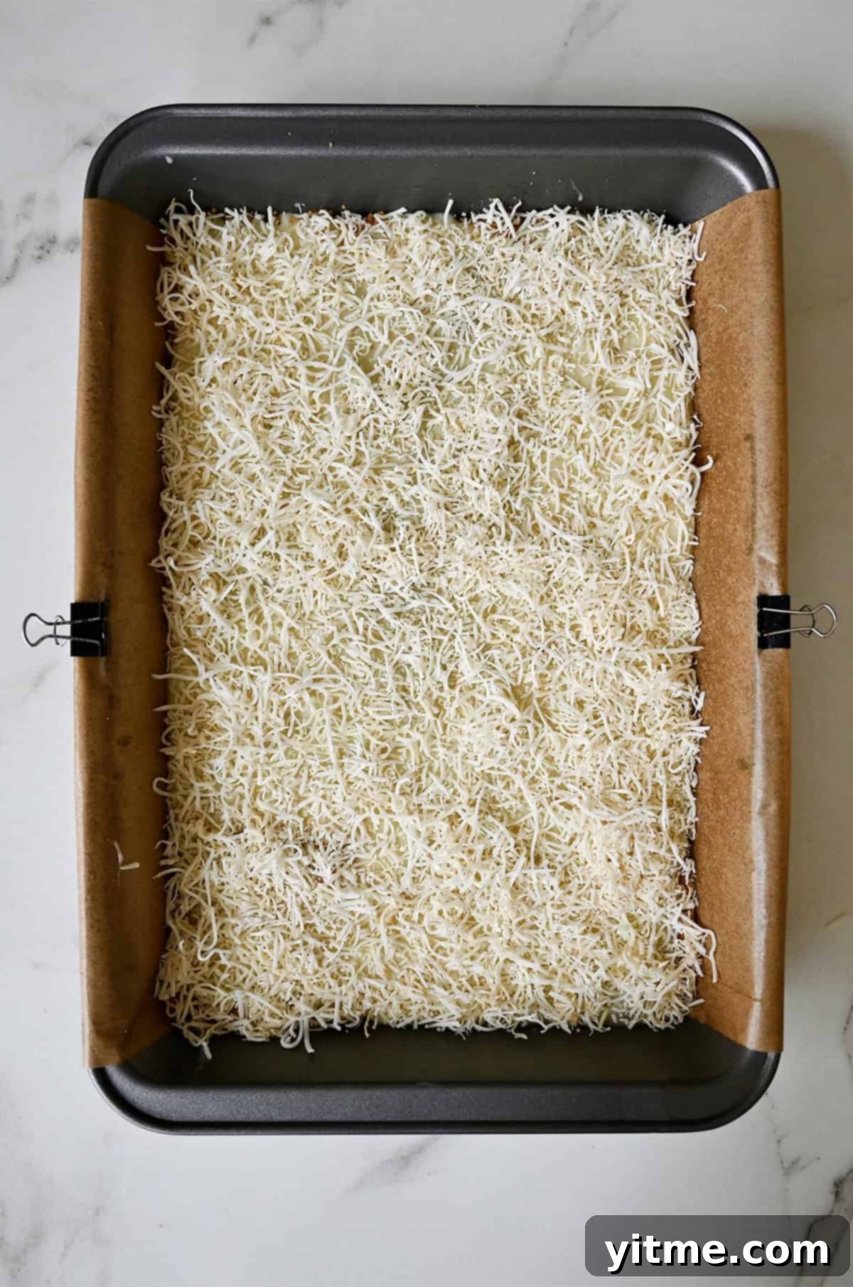 Everything Cookie Bars 10 Shredded coconut atop a layer of sweetened condensed milk and a graham cracker base in a parchment paper-lined pan, the first layer of toppings.