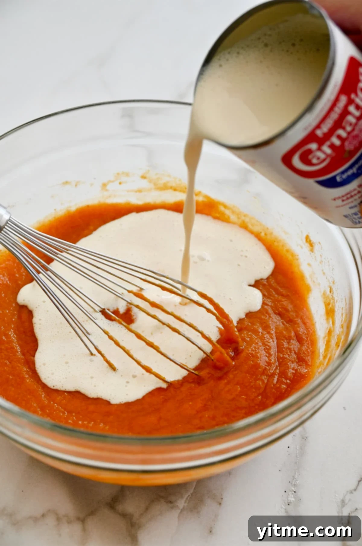 Golden Pumpkin Perfection 7 A hand whisking evaporated milk into a vibrant orange mixture of pumpkin puree, spices, and eggs in a clear glass bowl, preparing the pumpkin pie filling.