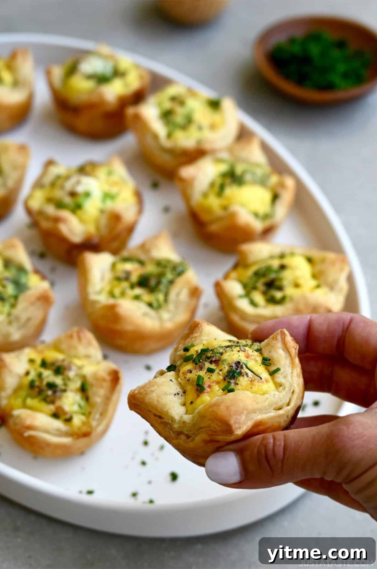 A hand holds a golden-brown mini quiche with a flaky puff pastry crust, ready to be enjoyed.
