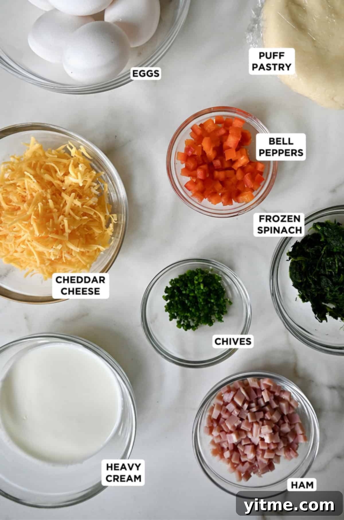 Various ingredients for mini quiches, including puff pastry, eggs, heavy cream, chives, and different fillings, neatly arranged in glass bowls.